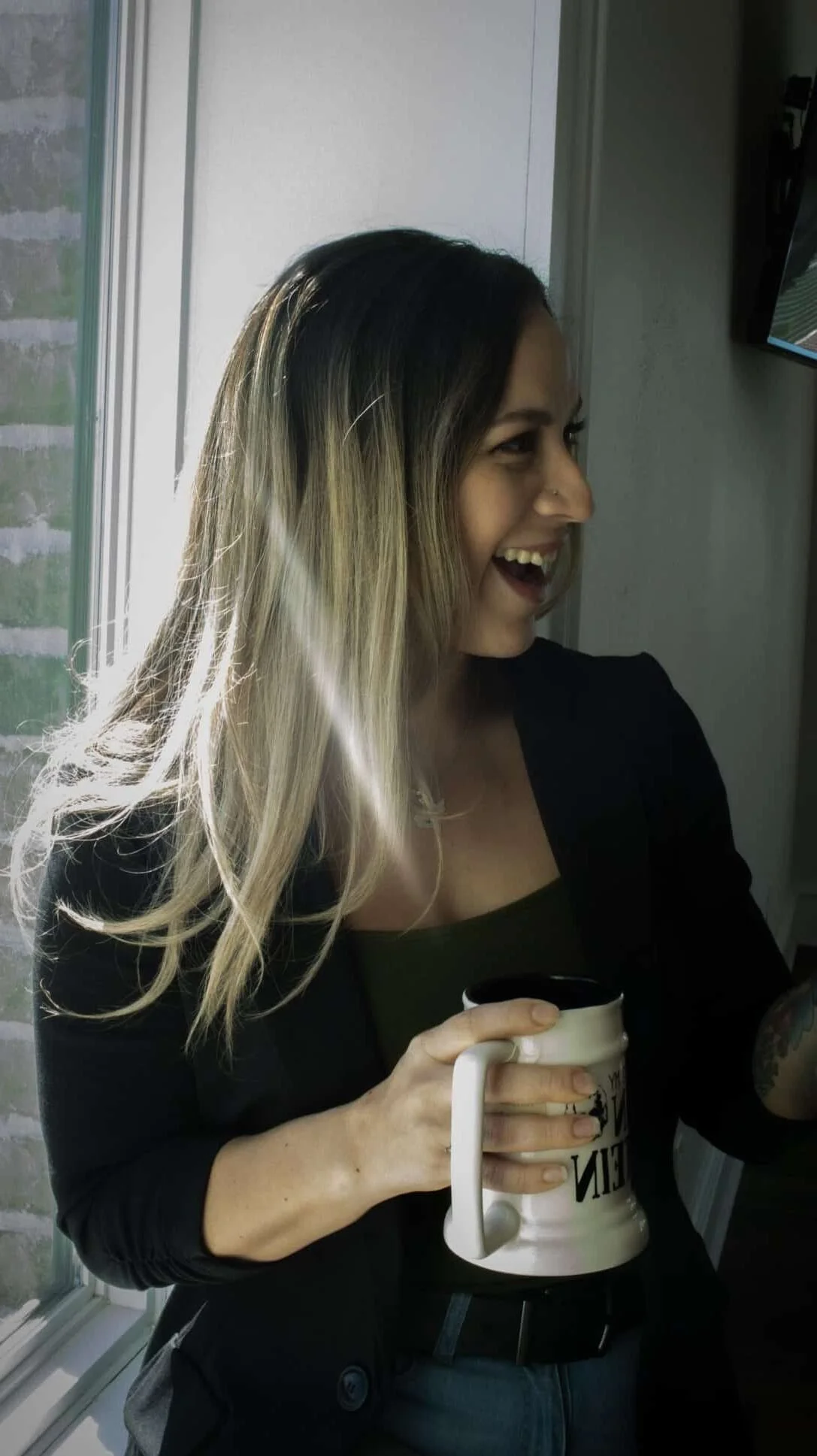 A woman with shoulder-length, ombre hair smiling and holding a coffee mug, standing near a window with sunlight shining through.