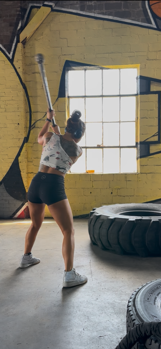 A woman in athletic clothing, wearing headphones, is swinging a sledgehammer at a tire in a gym with yellow brick walls and large windows.