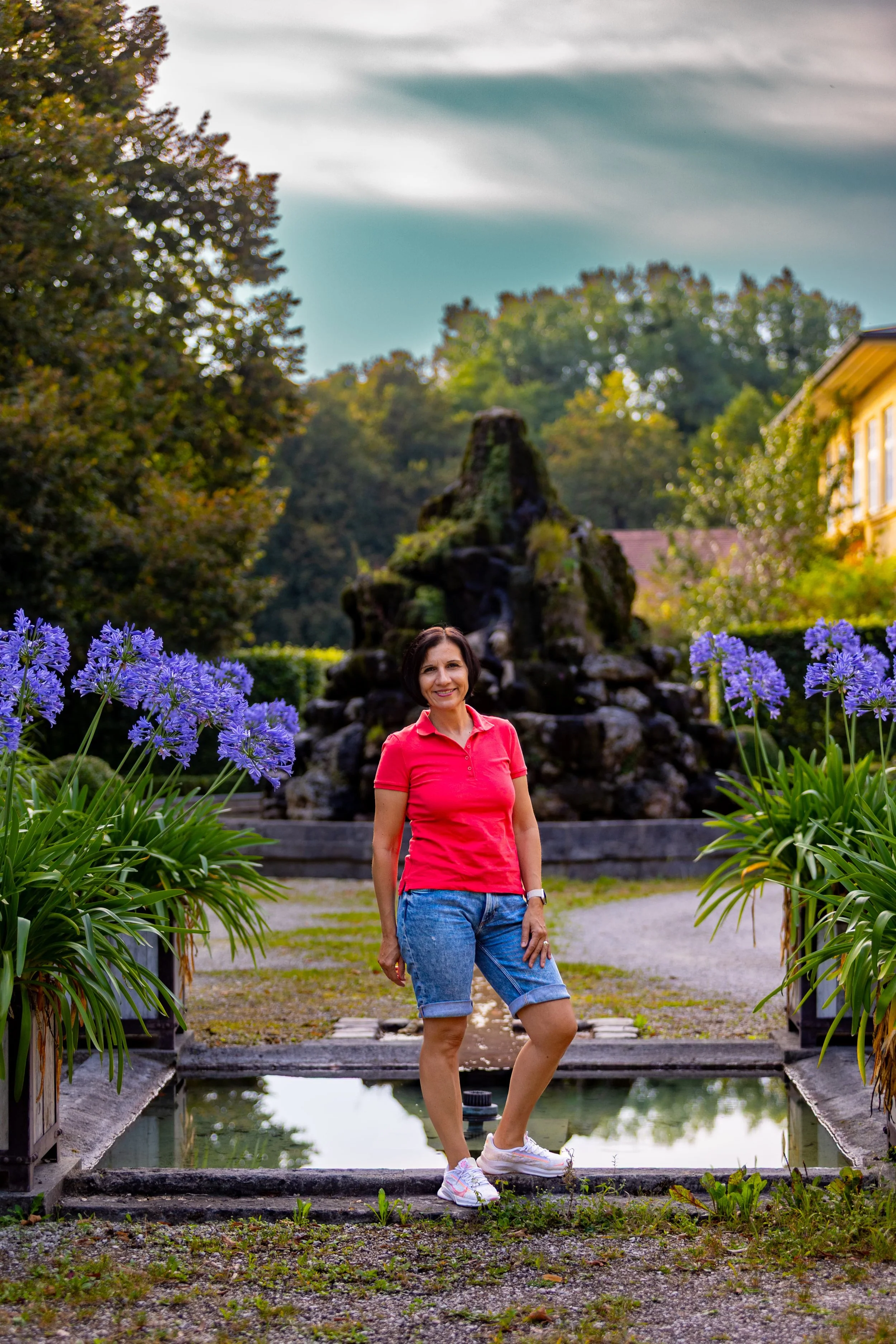 Clara Frankenberger steht in einem Garten vor einem Springbrunnen mit blauer Blume und einem großen Wasserfall im Hintergrund.