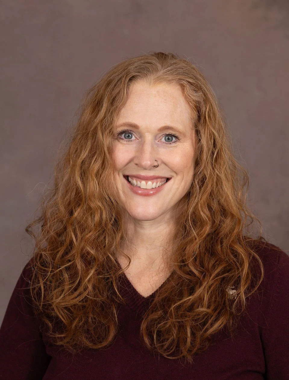 Josephine Eades smiling with curly red hair wearing a dark maroon top against a gray background.