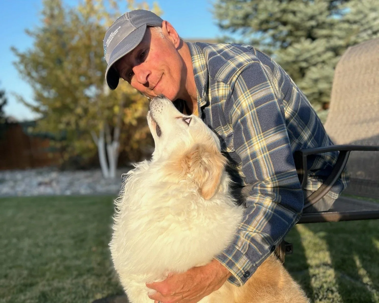 Dr. Dave Jackson in a plaid shirt and gray cap sitting outdoors, lovingly holding a Aussie (Eddie) as they touch noses, with trees and grass in the background.