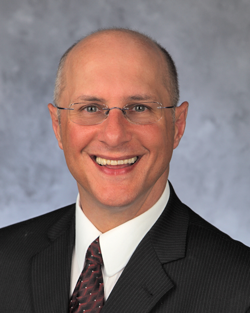 Professional headshot of a smiling man with glasses, wearing a dark suit, white shirt, and a patterned tie, against a neutral background.