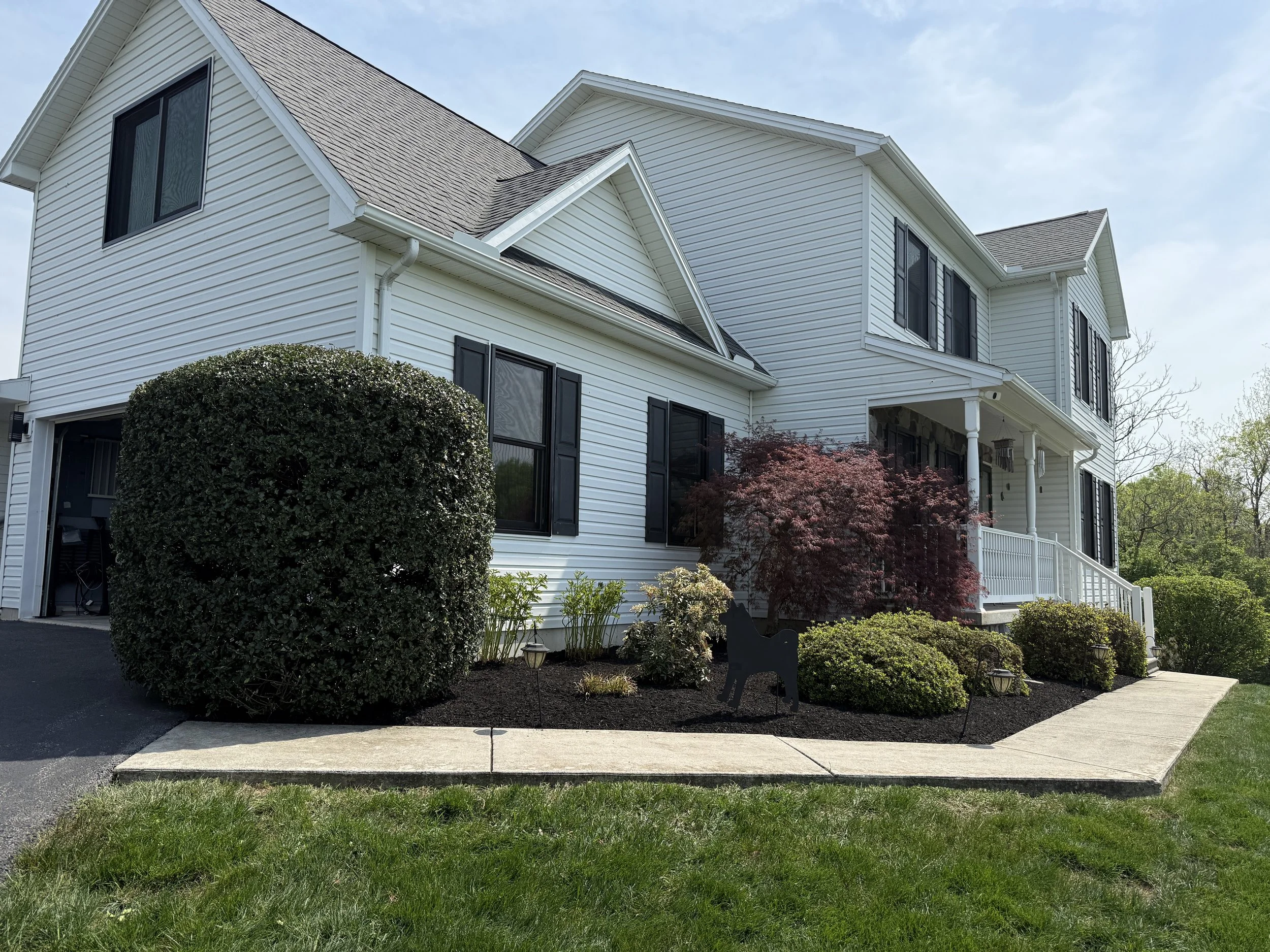 Front view of a white, two-story house with black shutters, a small porch with white railing, landscaped garden with bushes and a small red tree, sidewalk, and driveway on the left, under a partly cloudy sky.