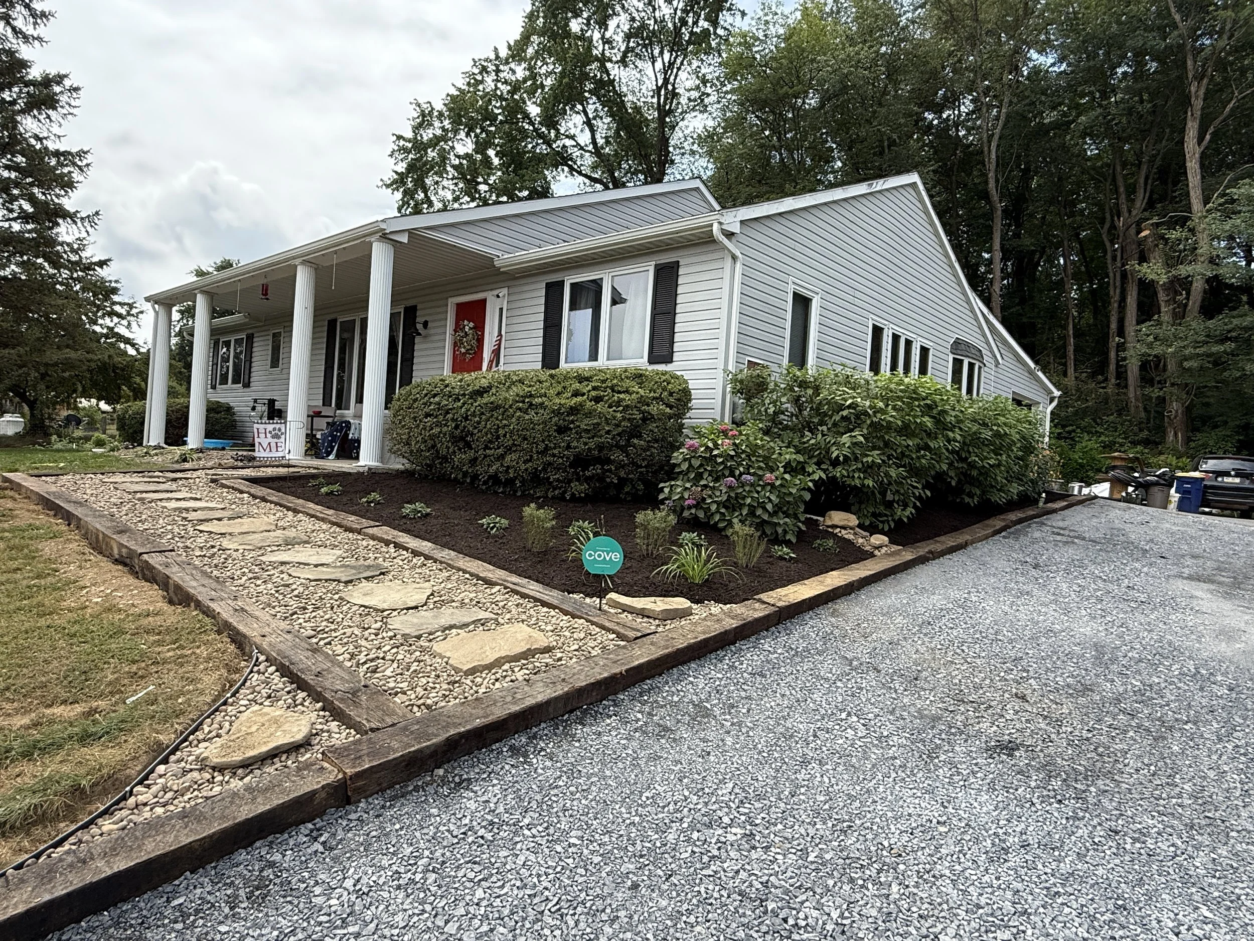 White house with black shutters and a red front door, surrounded by shrubs and a garden, with a gravel driveway in front.