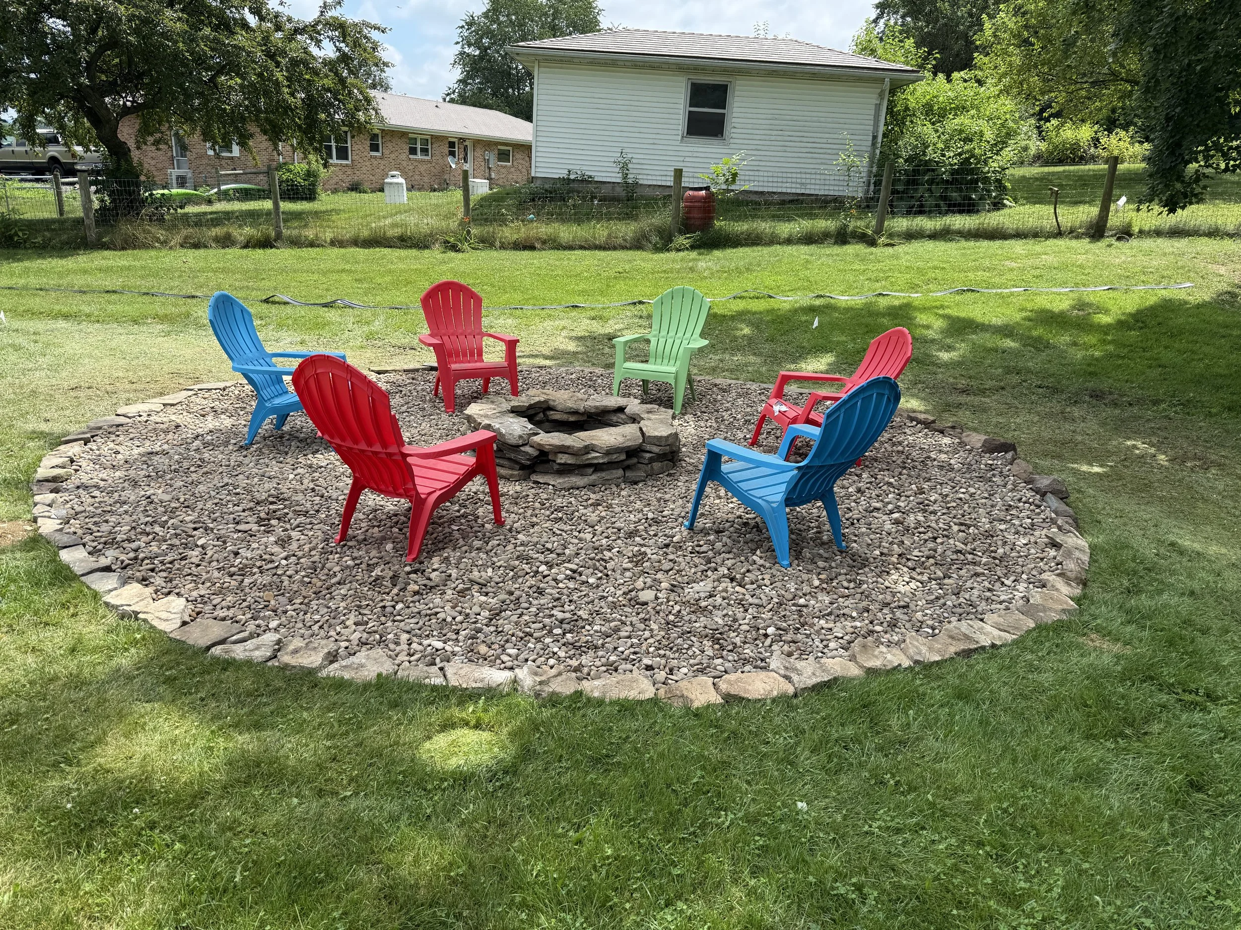 Colorful Adirondack chairs arranged in a circle around a stone fire pit on a gravel surface in a backyard.
