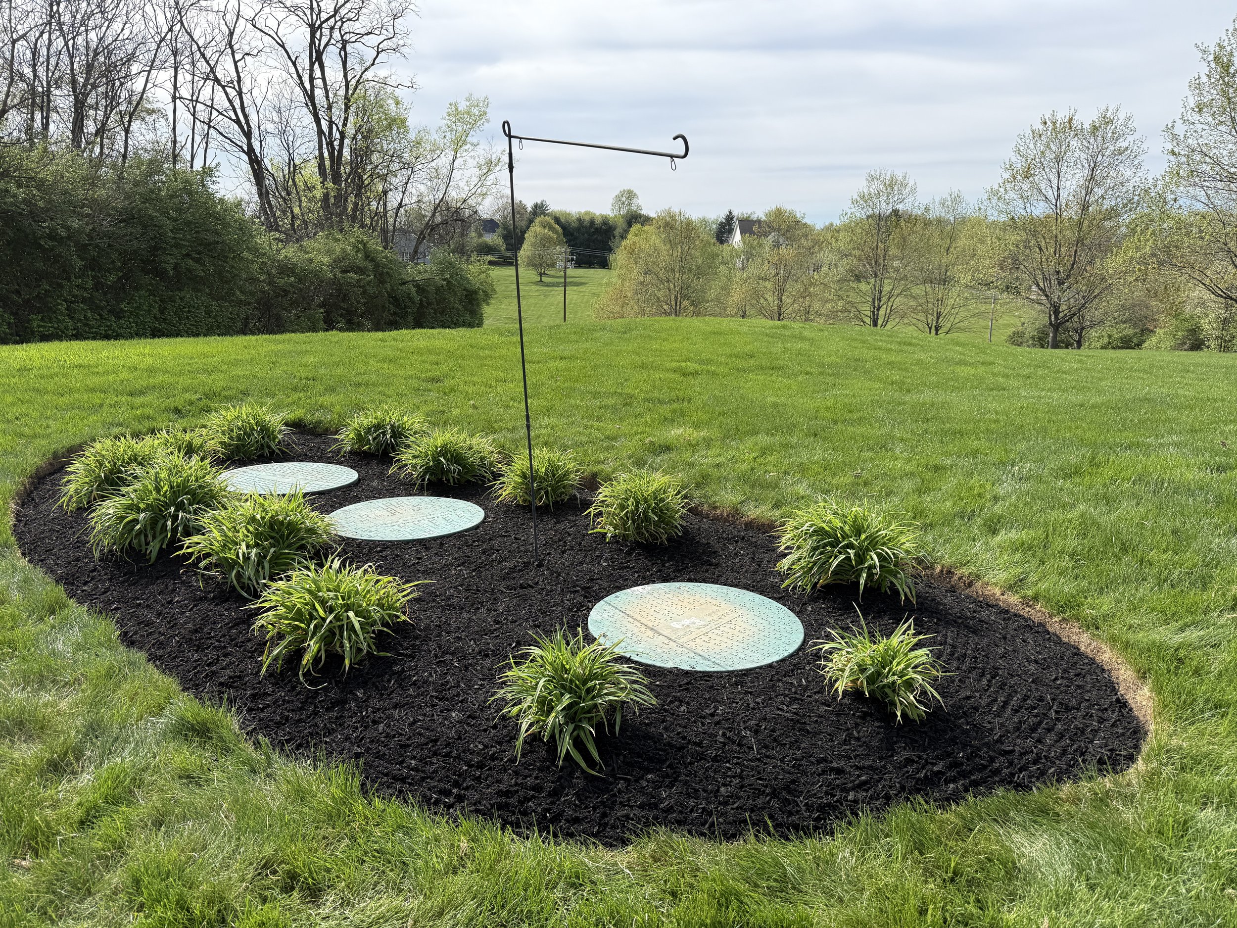 Well-maintained lawn with three circular underground utility access covers surrounded by ornamental grass, set in a landscaped garden bed on a grassy hill with trees in the distance under a cloudy sky.
