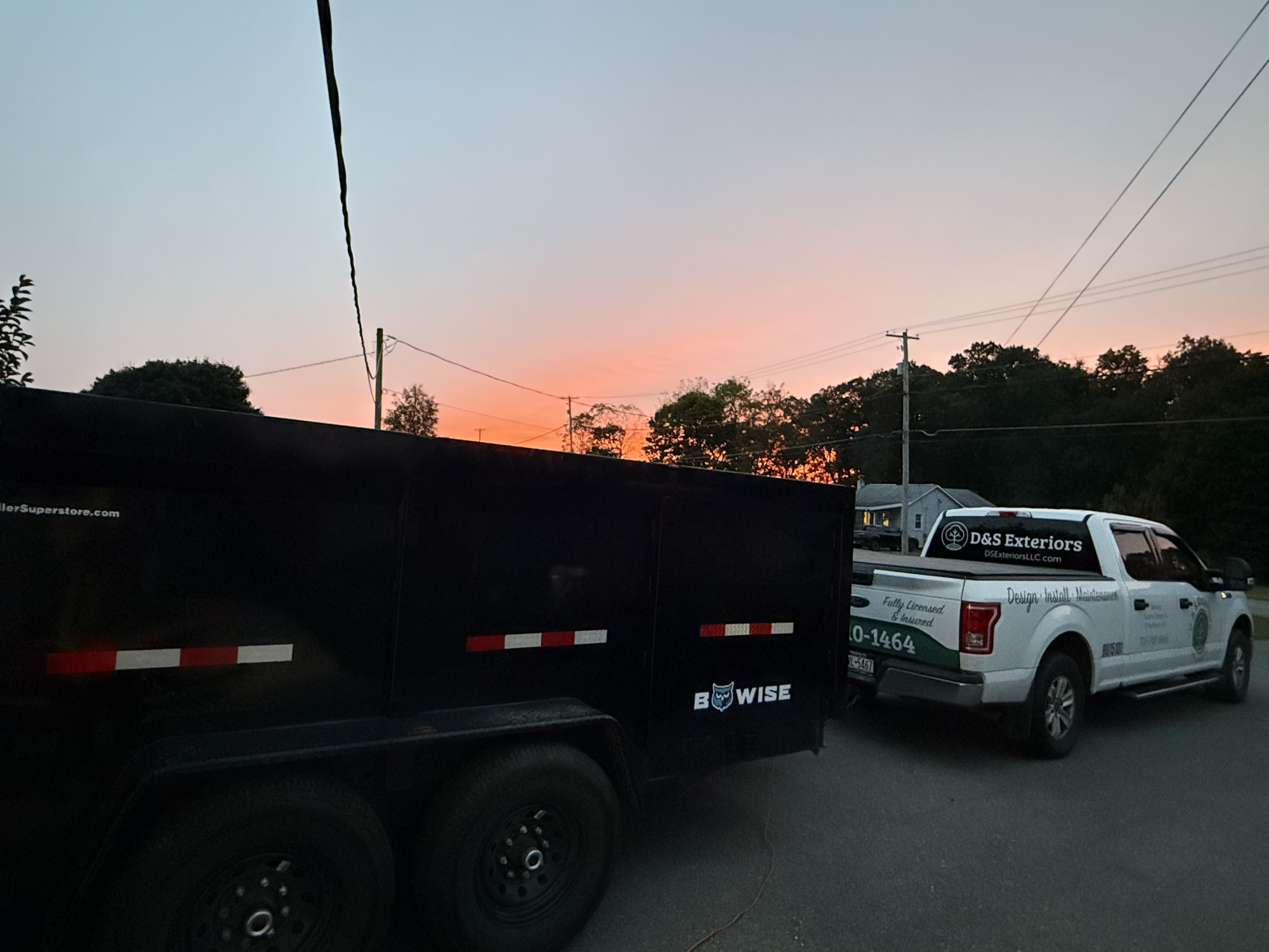 Vehicles parked on a street during sunset, including a black trailer with branding and a white work truck with company information.