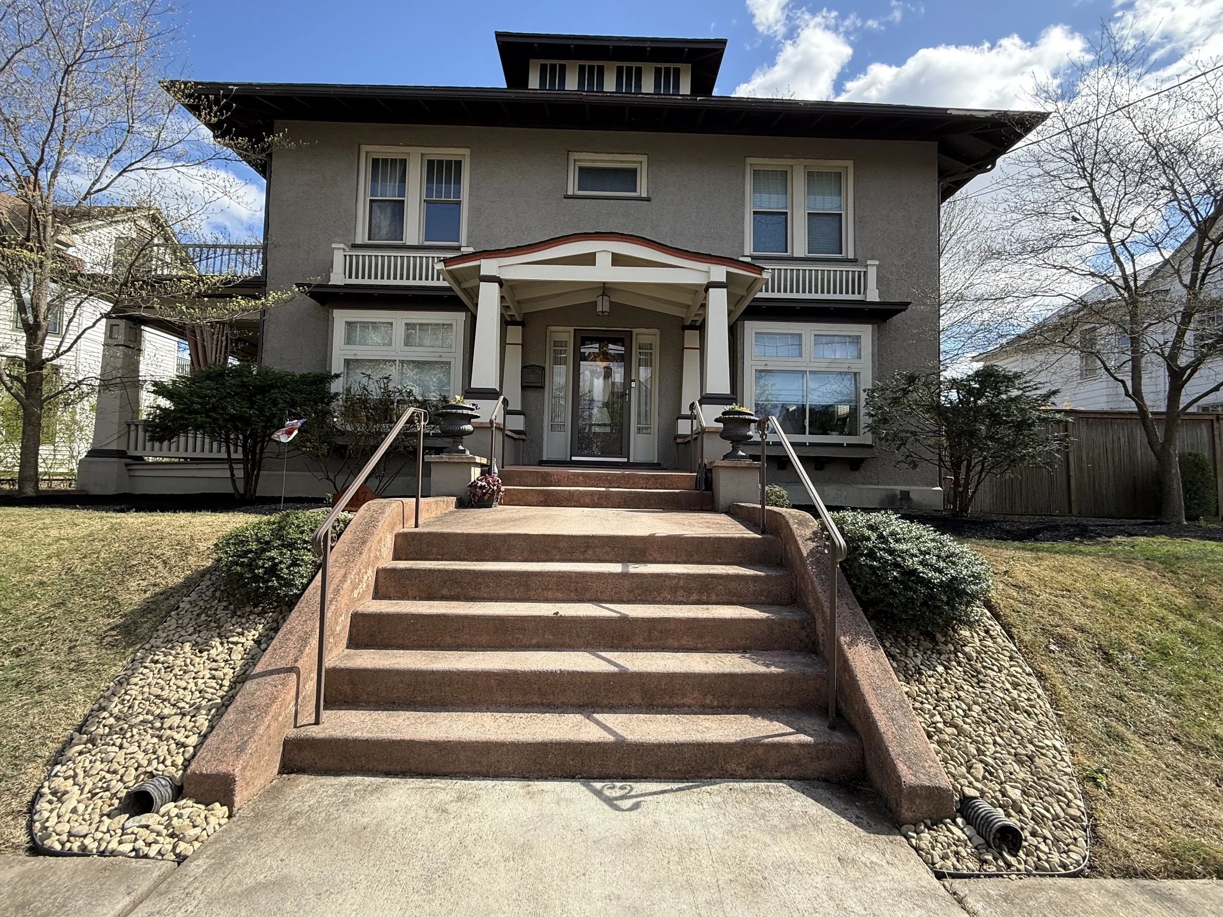A two-story house with a front porch, stairs, and landscaping, under a partly cloudy sky.