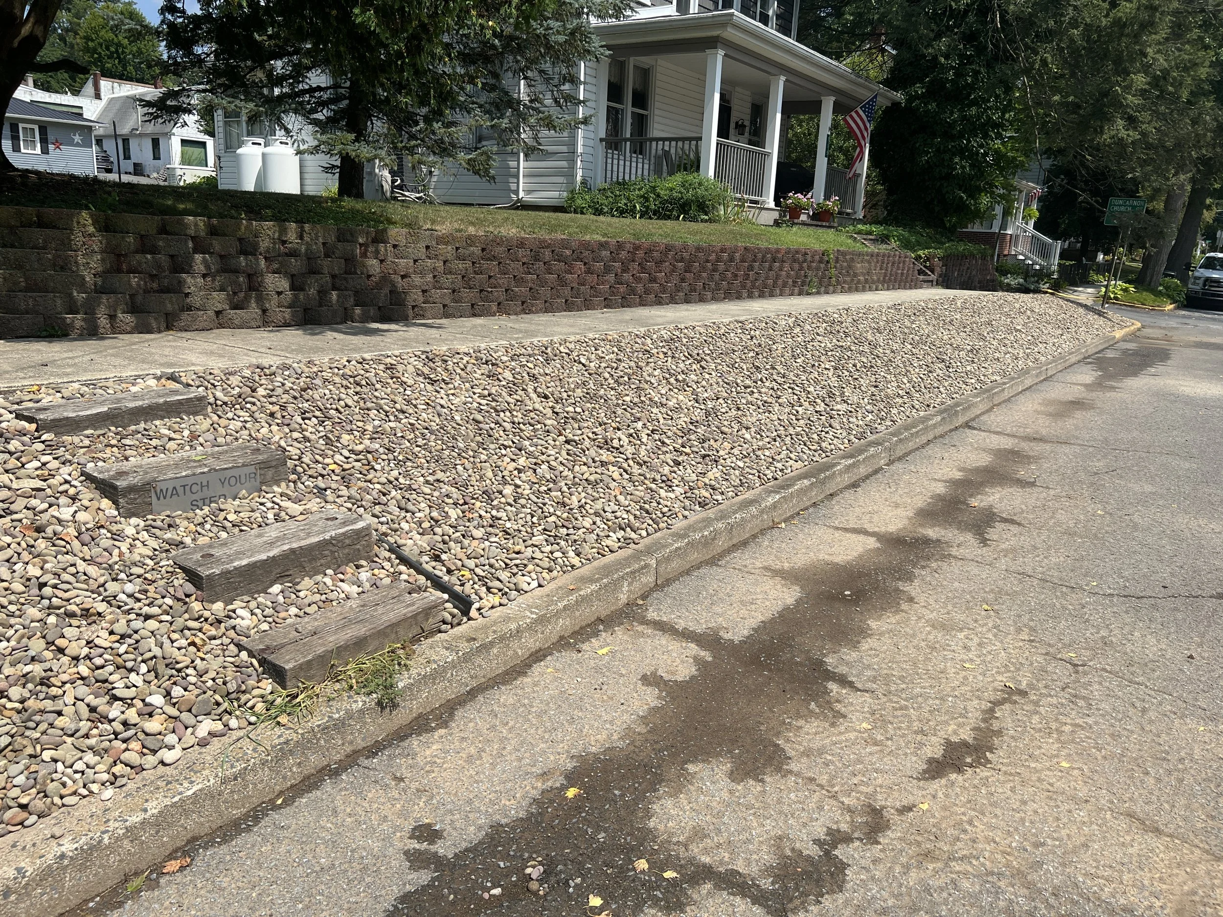 A residential street with a house featuring a porch with American flag, next to a gravelly yard with small rocks, weathered wooden steps, and a sign that reads 'Watch Your Step'.