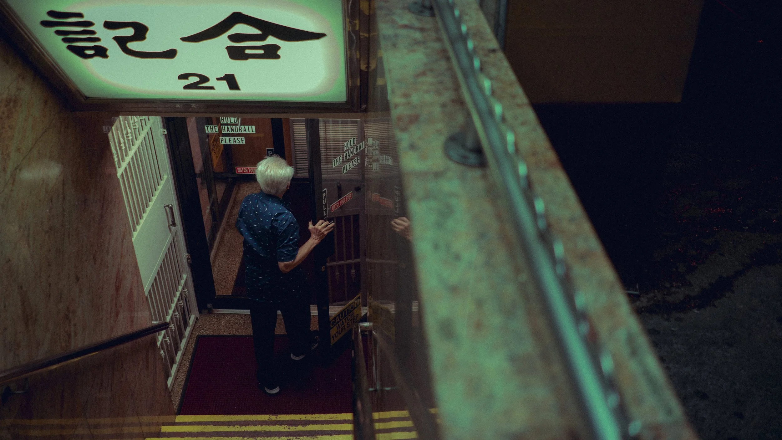 An elderly person with white hair, wearing a blue shirt with white dots, stands at the entrance of a building, opening the glass door.