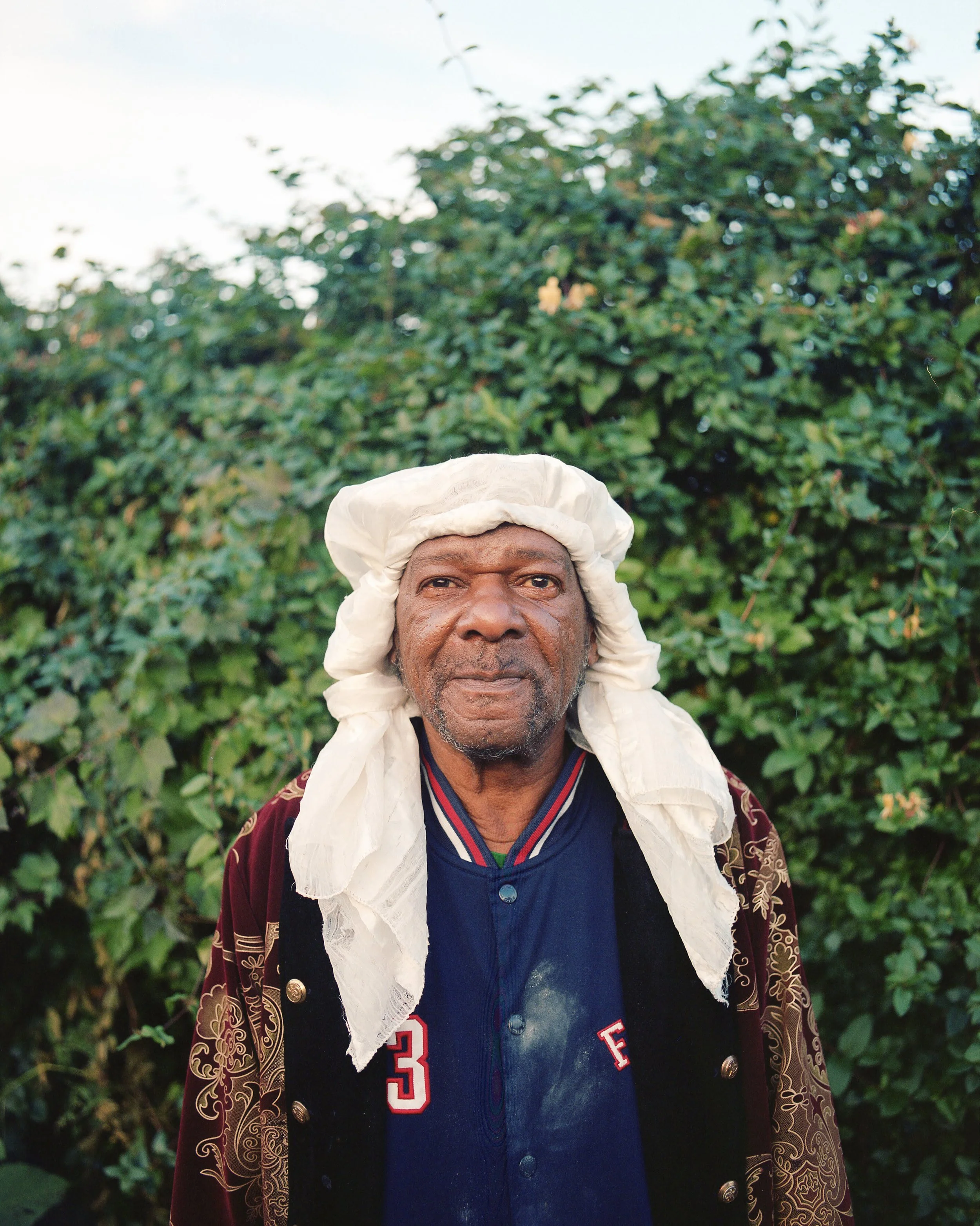 An elderly man wearing a white head covering, a navy blue shirt with red and white accents, and a patterned maroon jacket, standing outdoors in front of green bushes.
