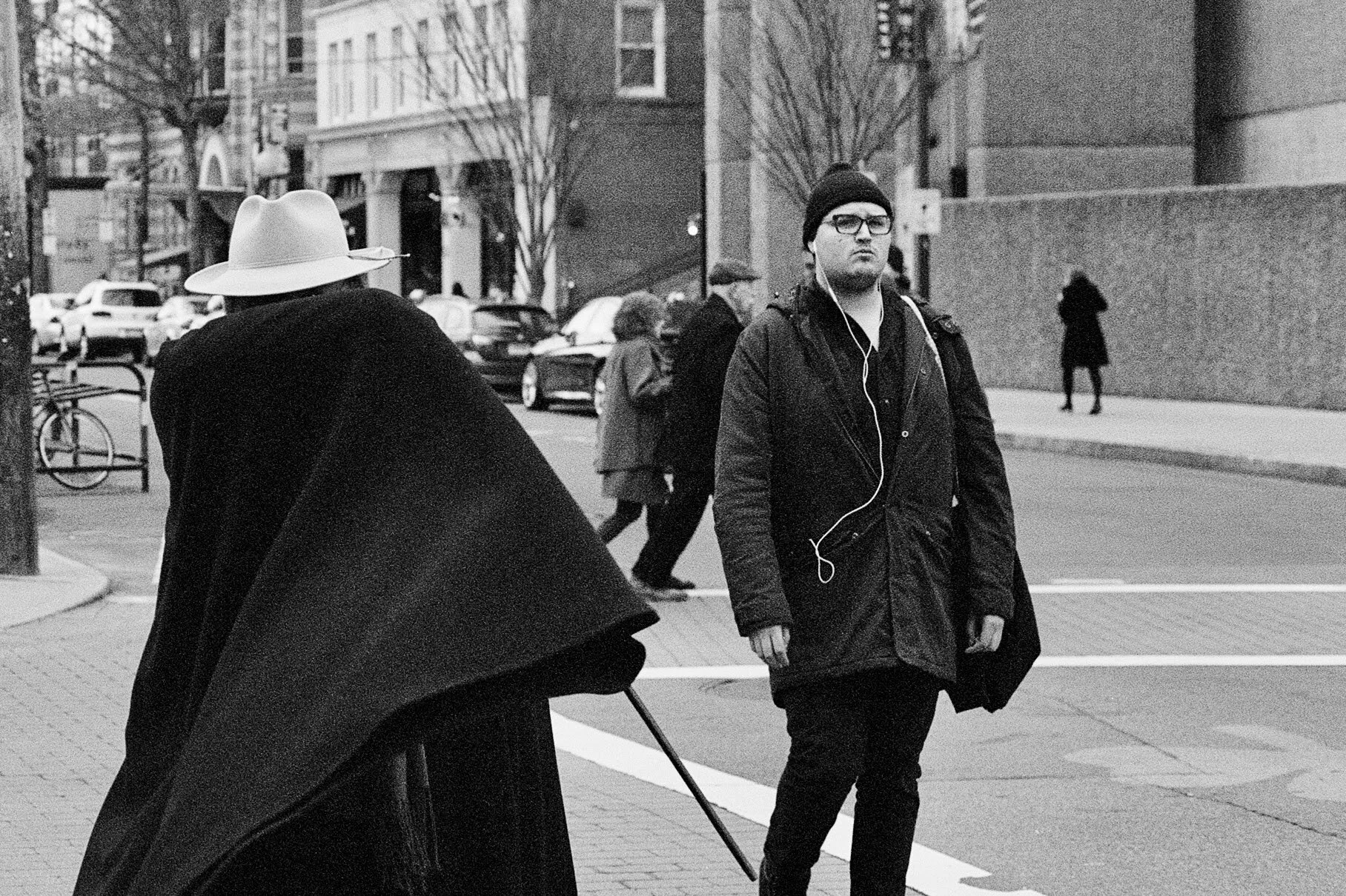 Black and white photo of a city street scene with pedestrians, including a man wearing glasses, headphones, and a beanie, walking past a person in a cape and wide-brimmed hat.