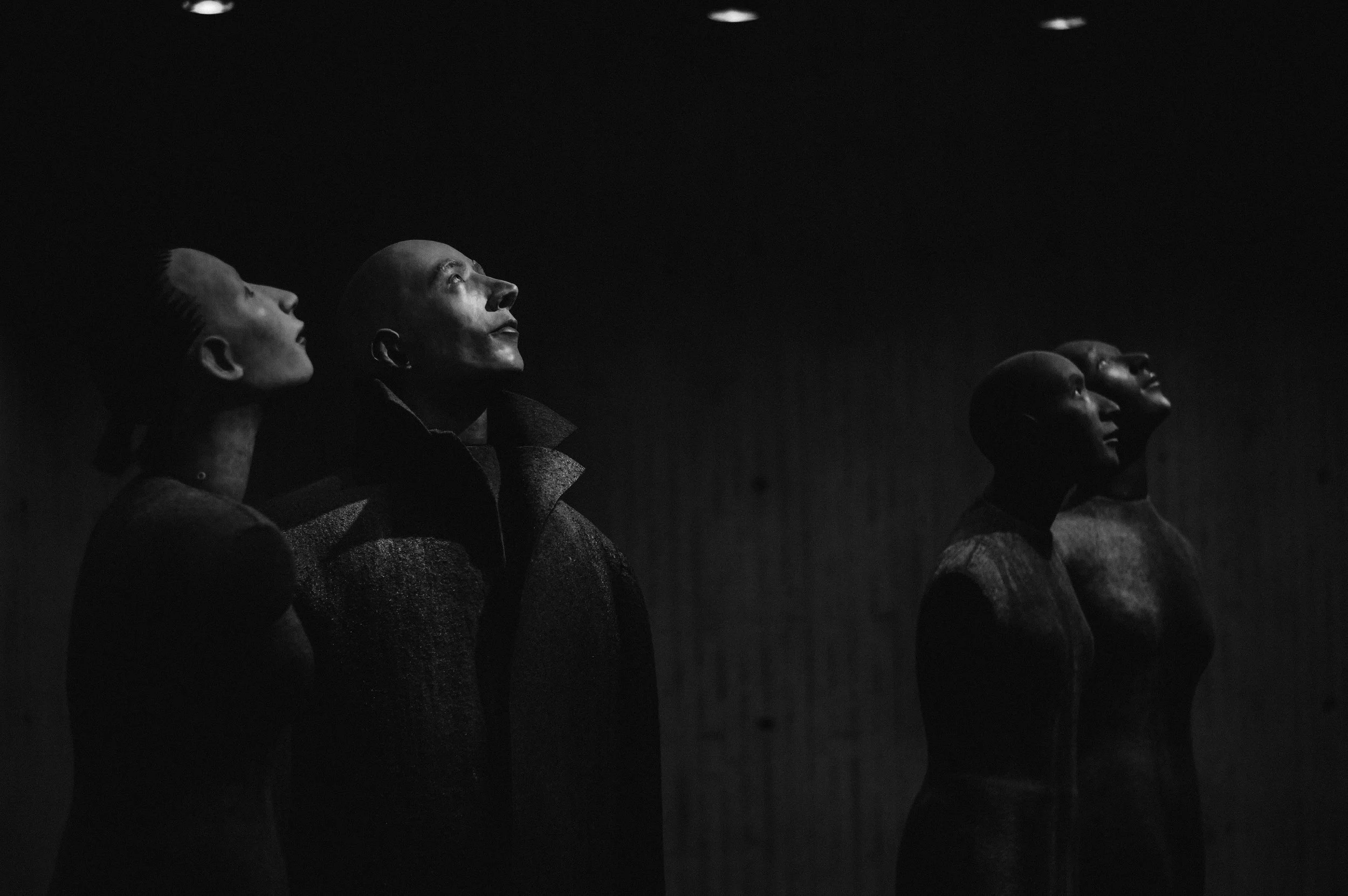 Four mannequins with bald heads and minimal facial features, standing in a dimly lit room, looking upward. They are dressed in various dark clothing.