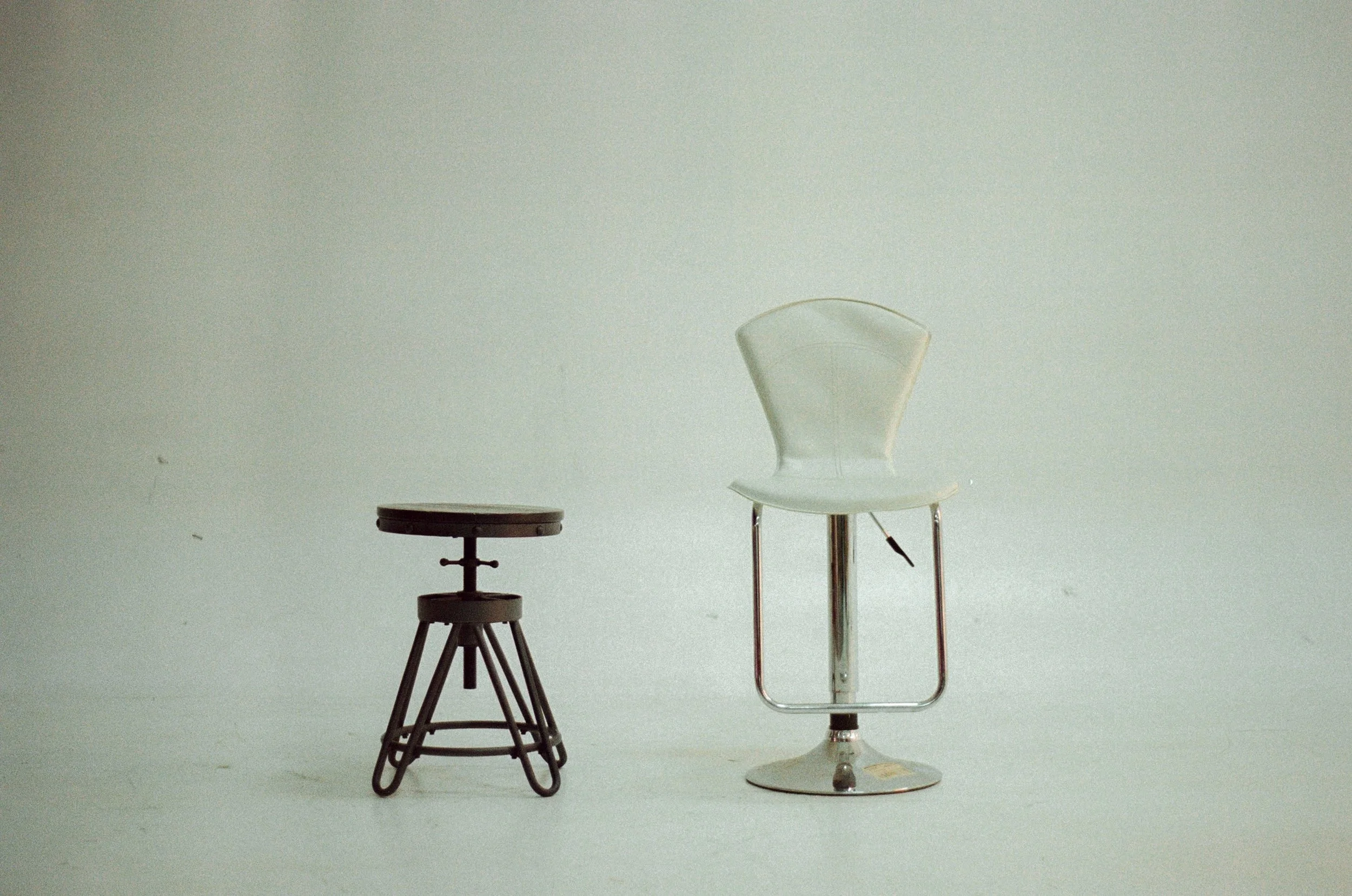 A black adjustable stool and a white modern office chair with a chrome base, set against a plain light-colored background.