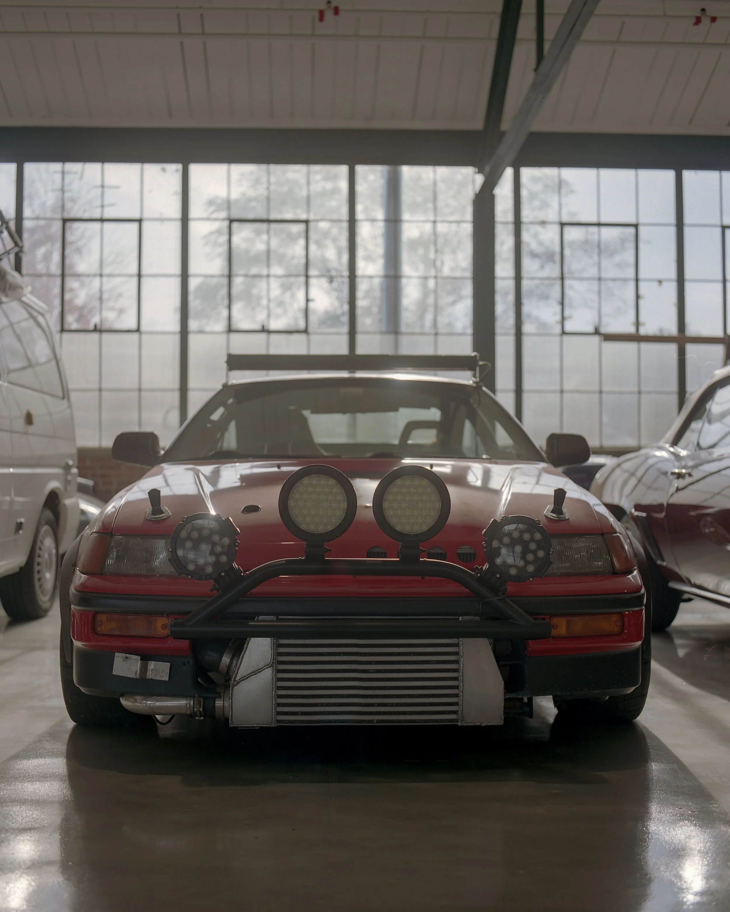 Red race car with large front-mounted radiator and additional lights, parked inside an industrial-style garage with large windows.