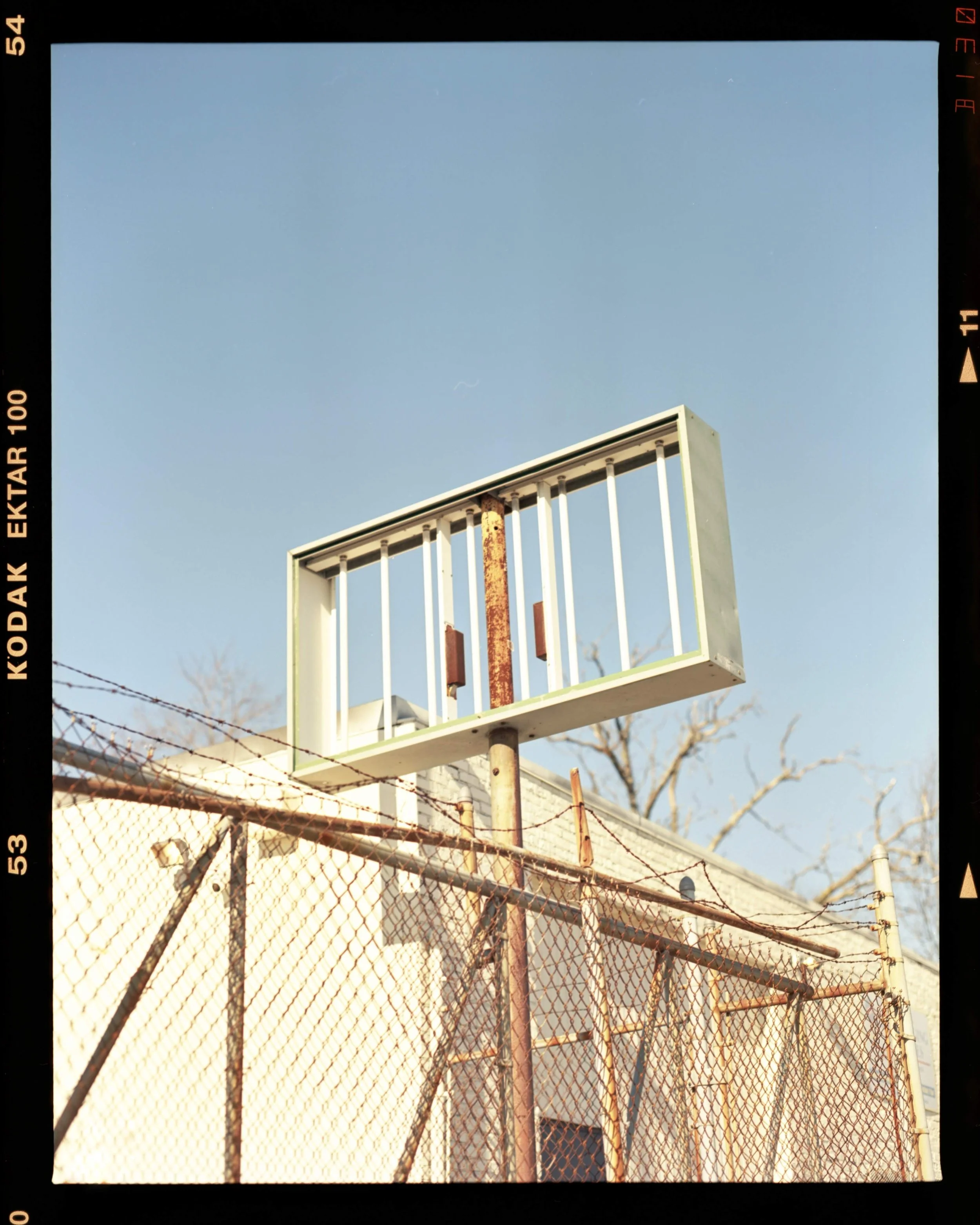 An empty abandoned basketball hoop with a rusted backboard frame, attached to a rusted pole, behind a chain-link fence under a clear blue sky.