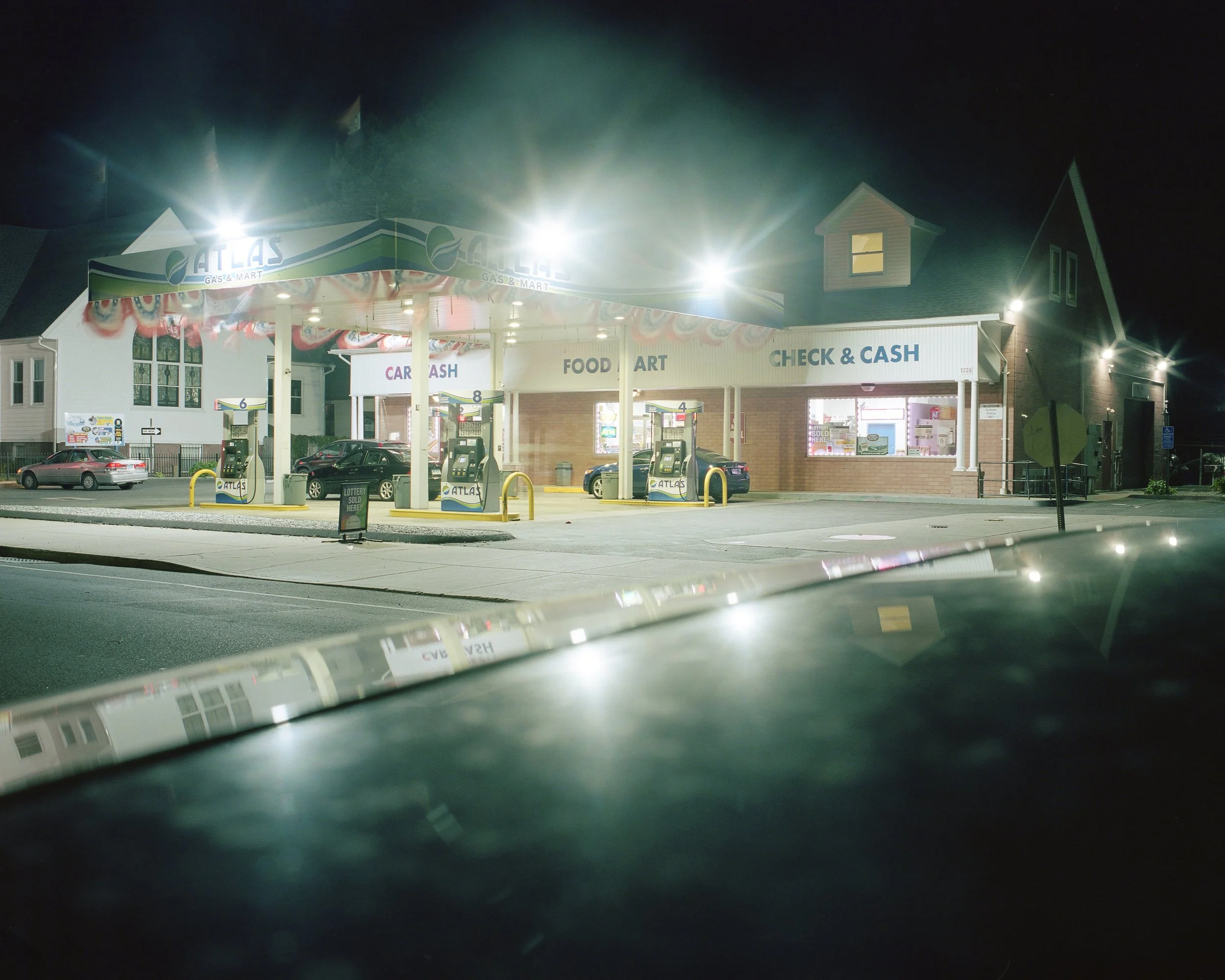 Night view of a gas station with multiple fuel pumps, a convenience store, and parked cars, illuminated by bright lights.