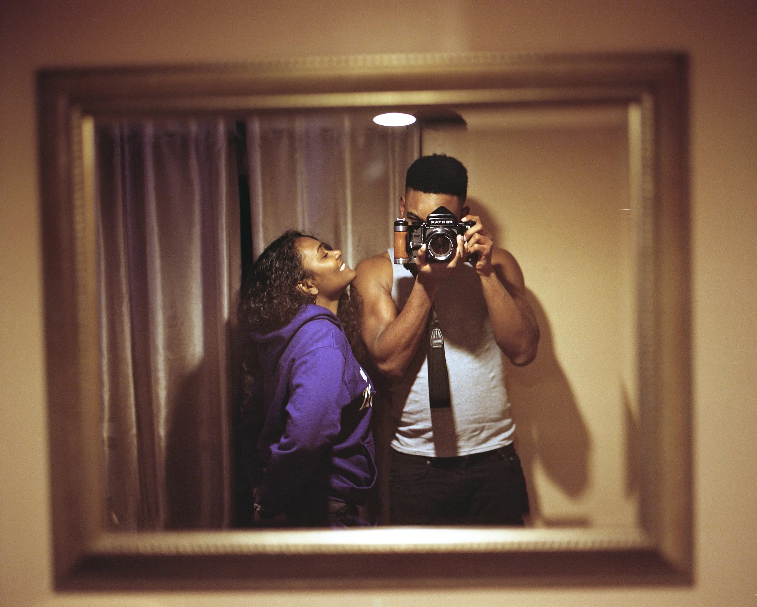 A woman and a man taking a selfie in a mirror. The woman is smiling and wearing a purple hoodie; the man is holding a camera and wearing a sleeveless shirt.