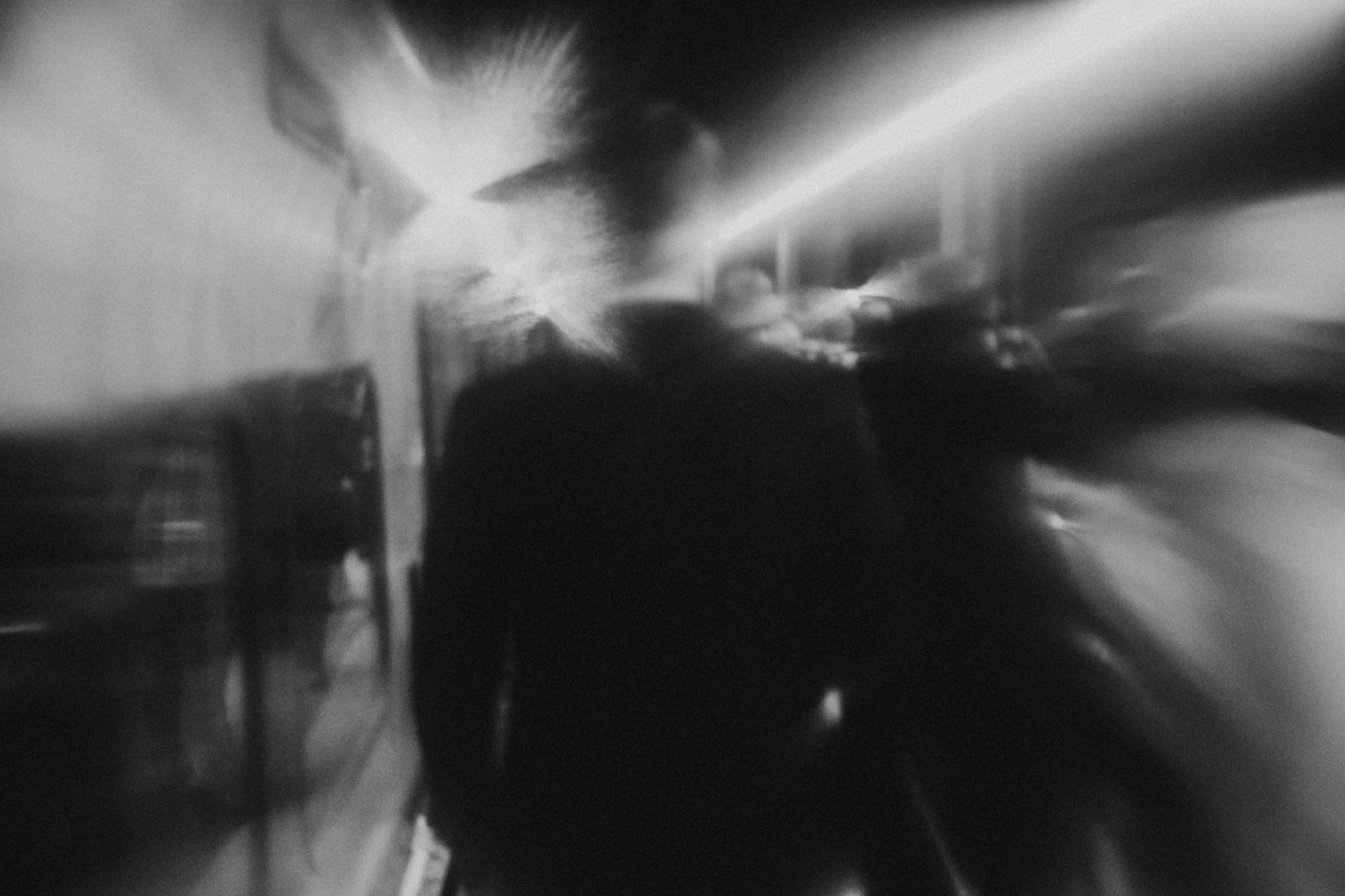 A blurry, black-and-white photo of two people walking in a corridor or hallway at night, with light streaks and abstract motion blur.