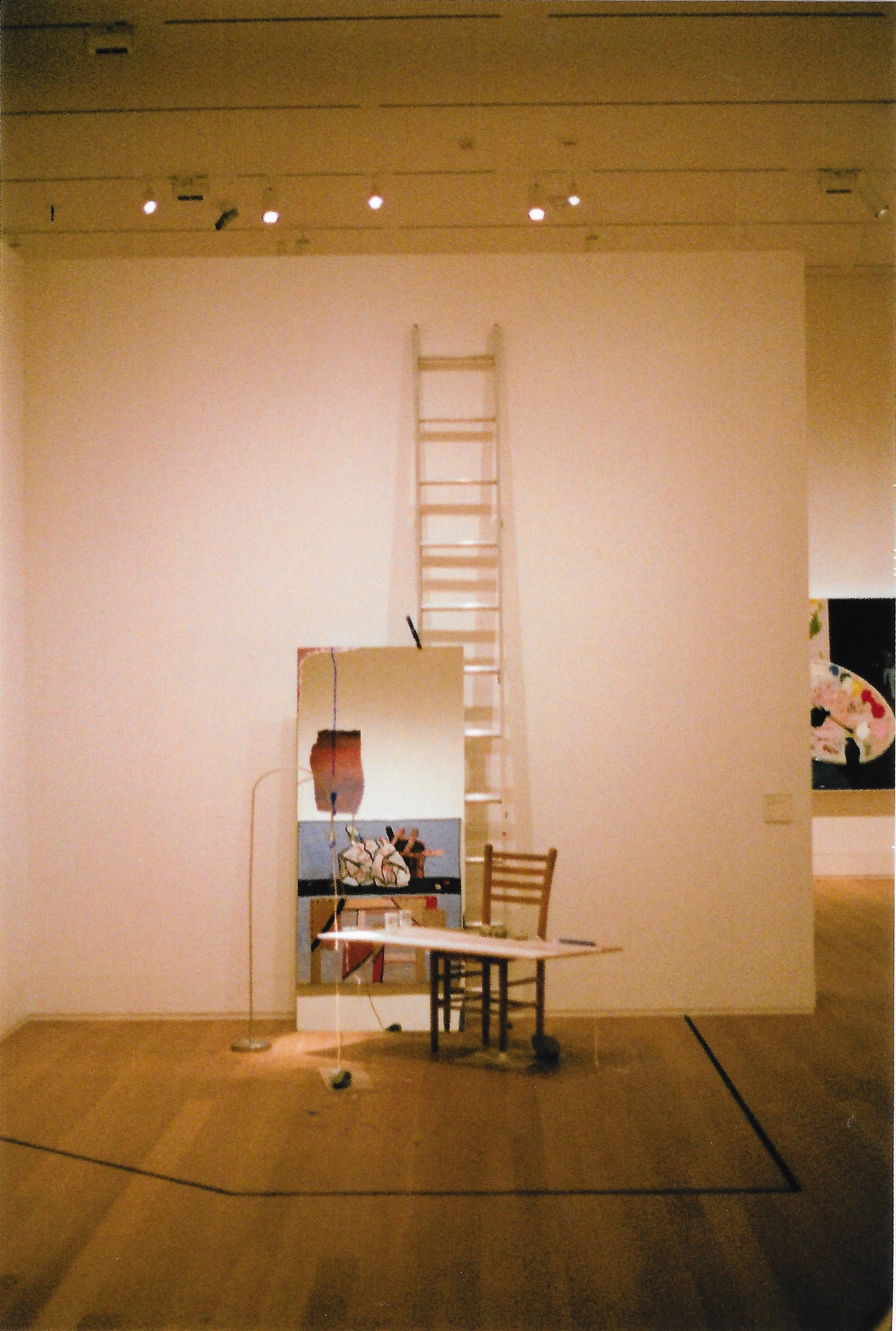 Minimalist art installation with a wooden ladder leaning against a white wall, a wooden chair, a tabletop with papers, a standing floor lamp, and artwork with abstract and floral designs in the background, all inside an art gallery room.