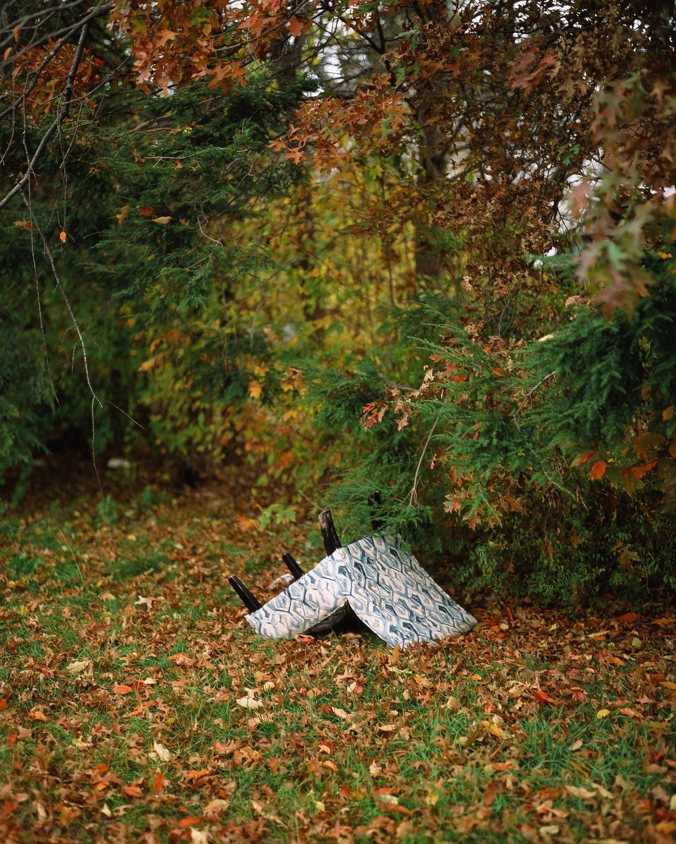 A discarded patterned tablecloth or fabric draped over an overturned picnic table in a wooded area with fall-colored leaves on the ground and on the trees.