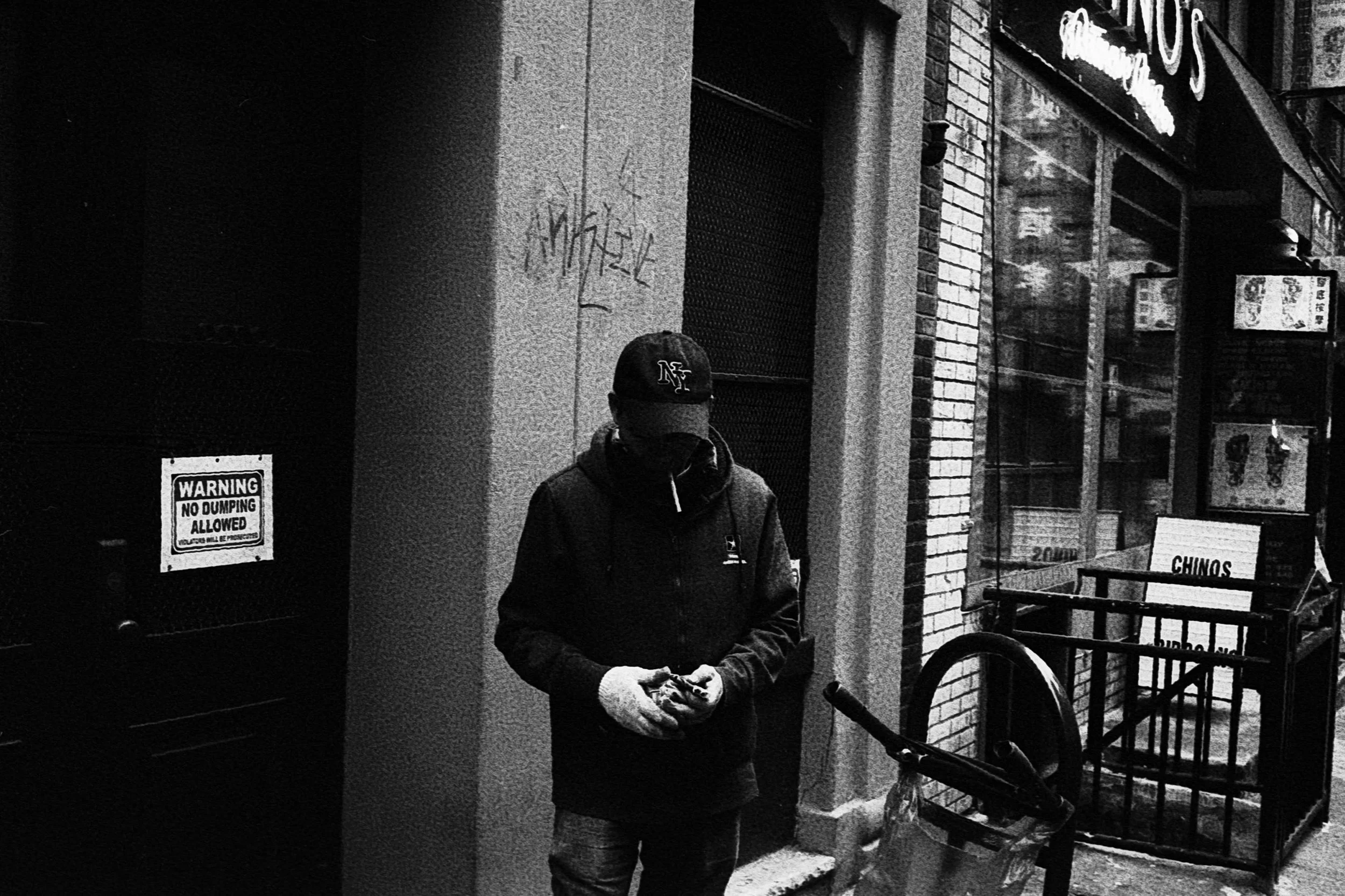 A man wearing a cap, jacket, and gloves standing on a city sidewalk looking at his phone. There is graffiti on the wall behind him, and a sign on a nearby door reads 'Warning No Dumping Allowed.'