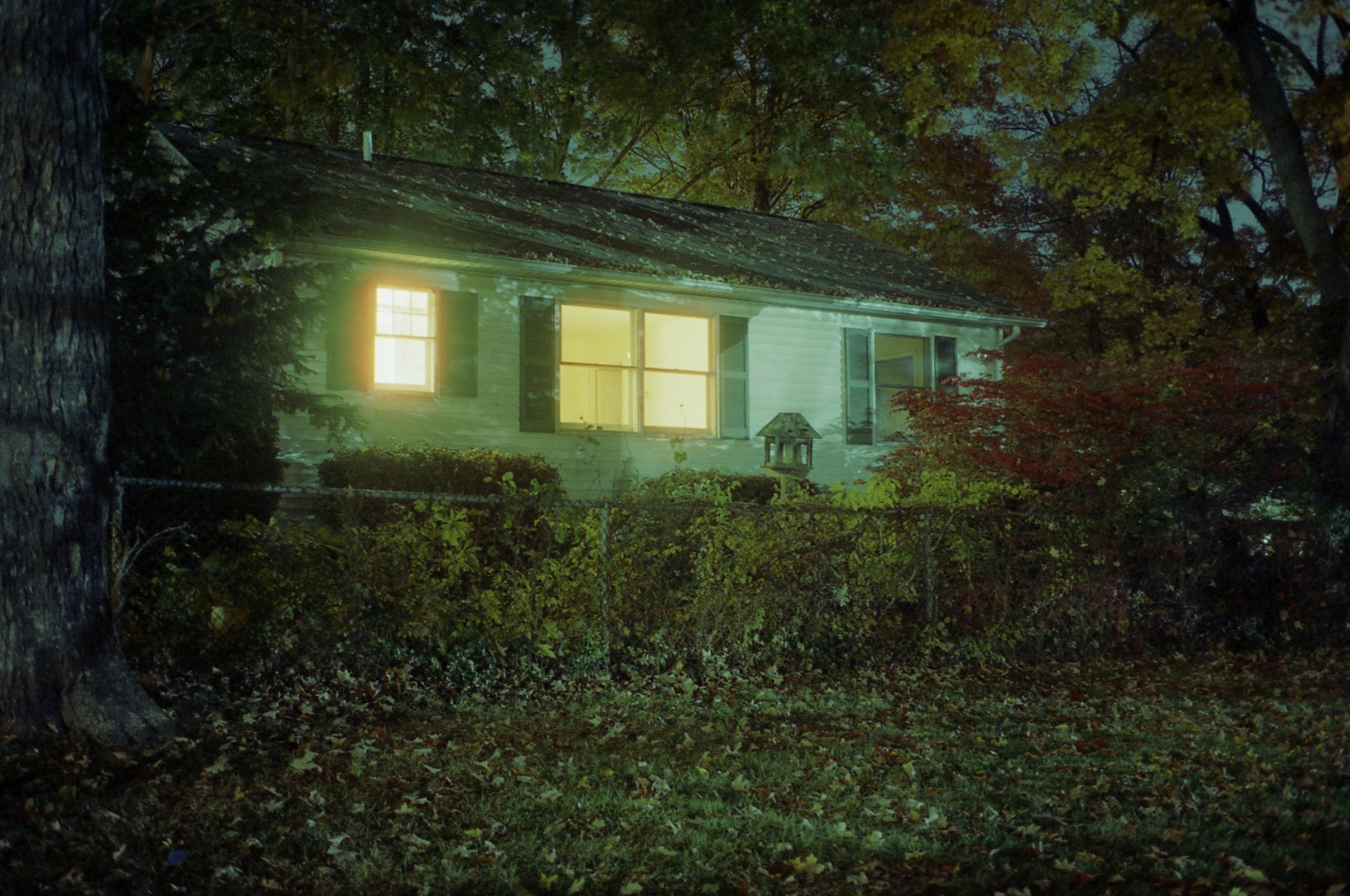 A house with illuminated windows at night, surrounded by trees and a fence, with leaves on the ground.