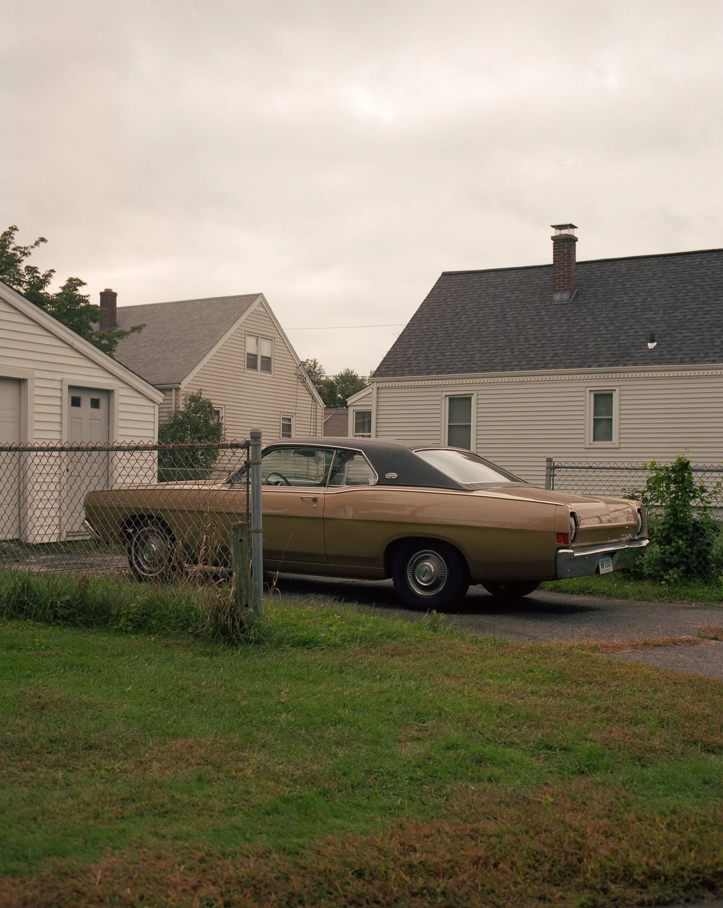 A vintage beige car parked on a driveway behind a chain-link fence, with two white houses and a cloudy sky in the background.