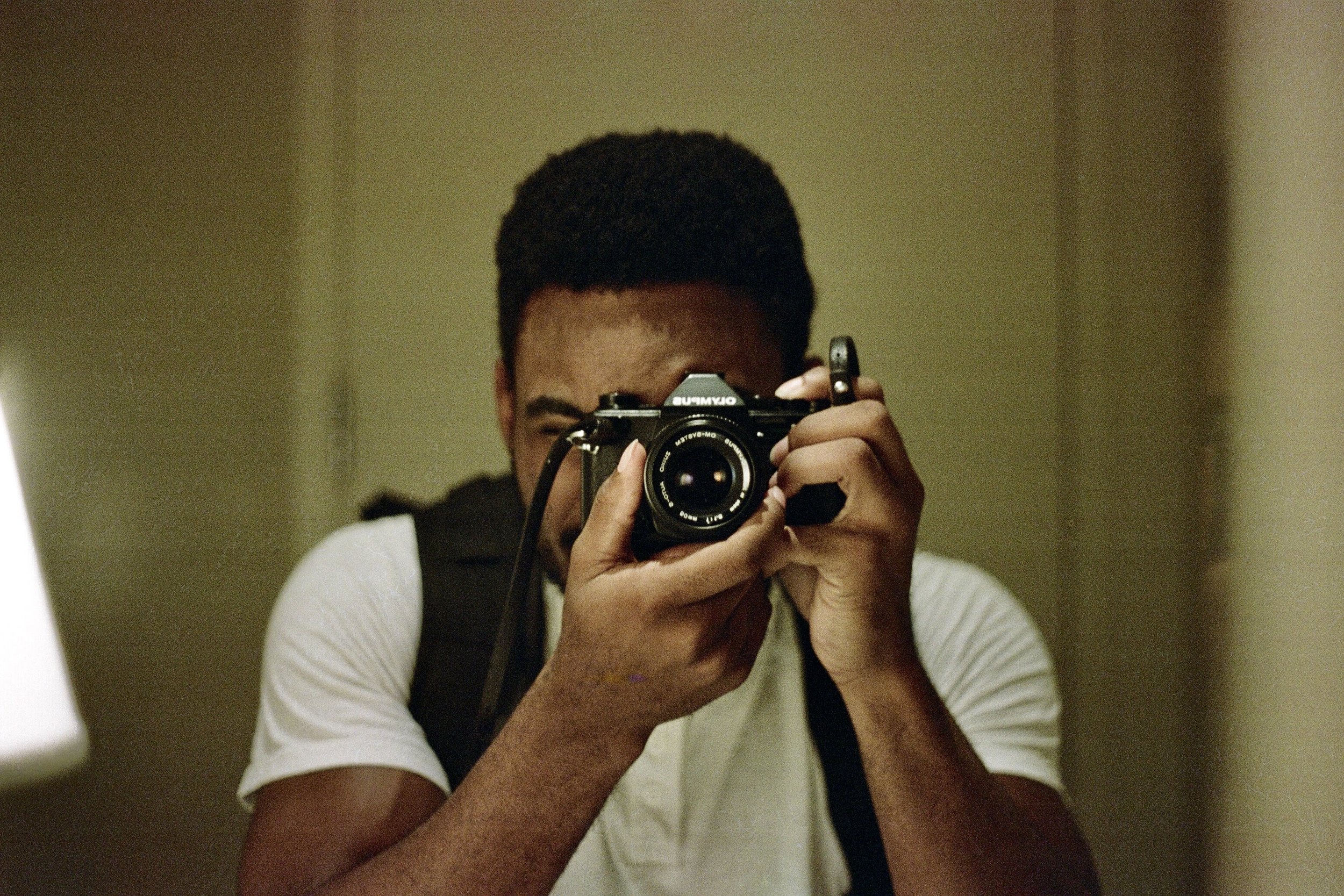 A young man with short curly hair taking a photo of himself with a compact camera in a mirror. He is wearing a white T-shirt and carries a black backpack.