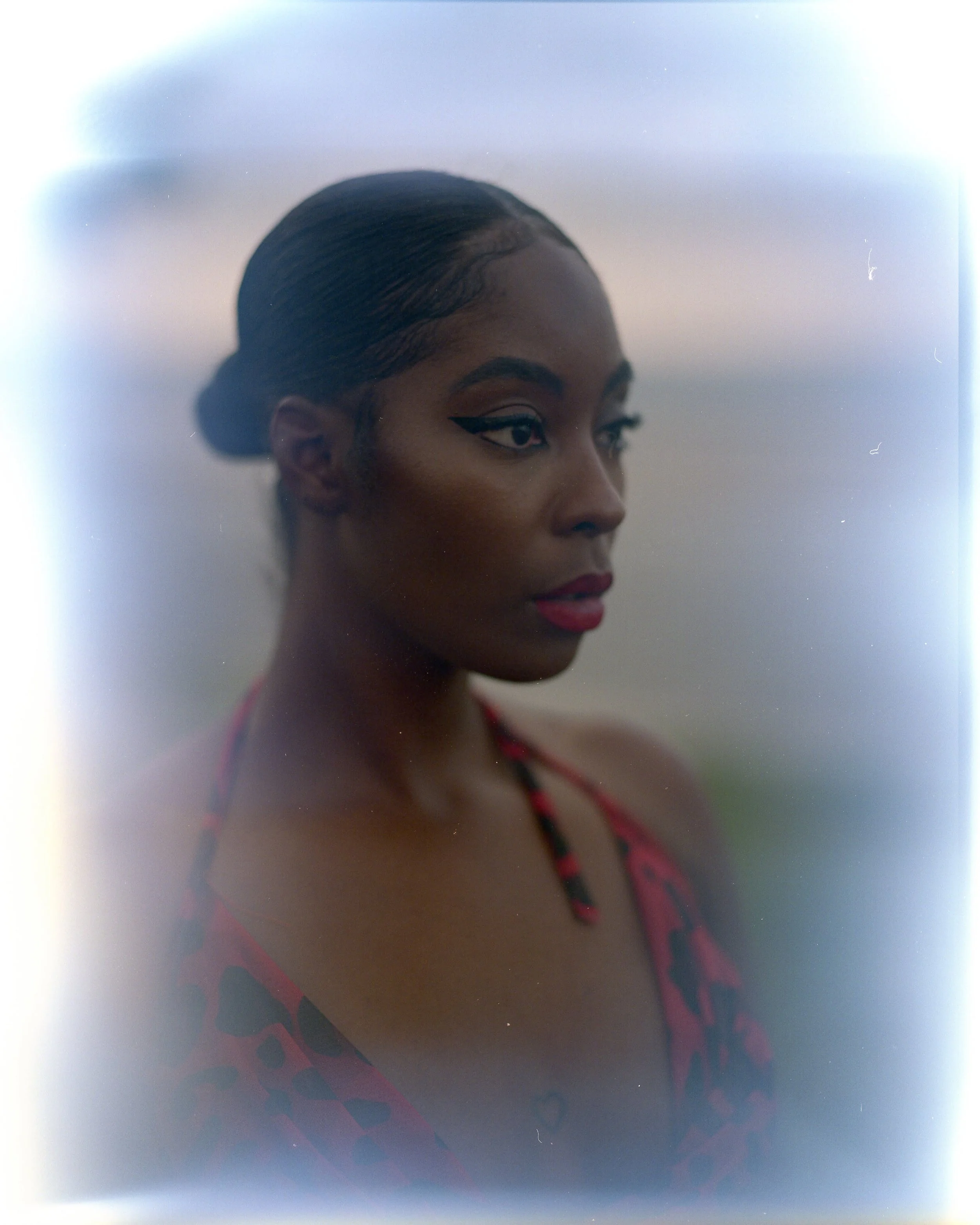 A woman with dark skin and styled hair, wearing makeup, including winged eyeliner and red lipstick, dressed in a red and black patterned top, looking to the side with a calm expression.