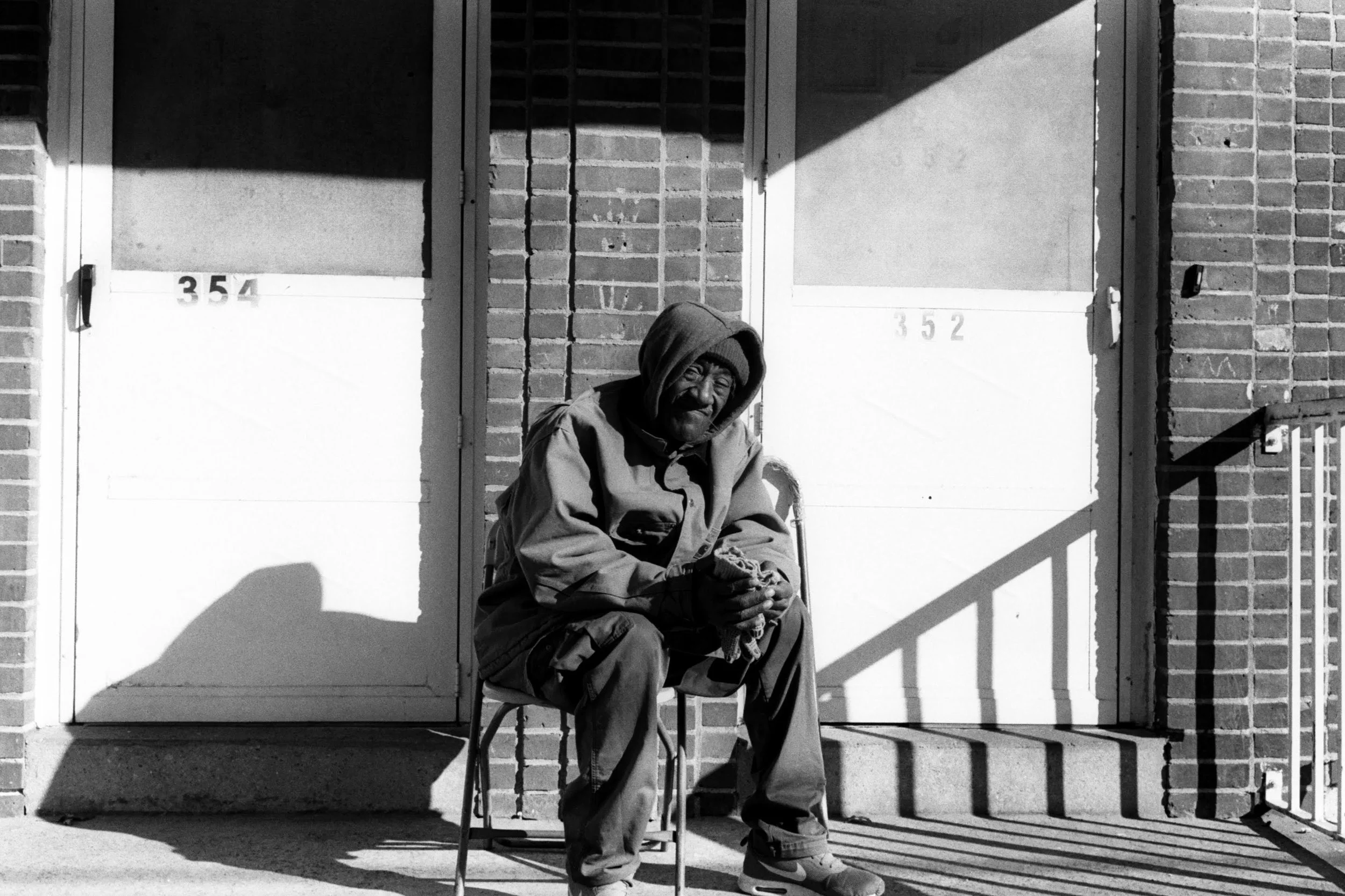 An elderly man sitting on a chair outside a building, wearing a hooded jacket, with shadows cast on the wall behind him.