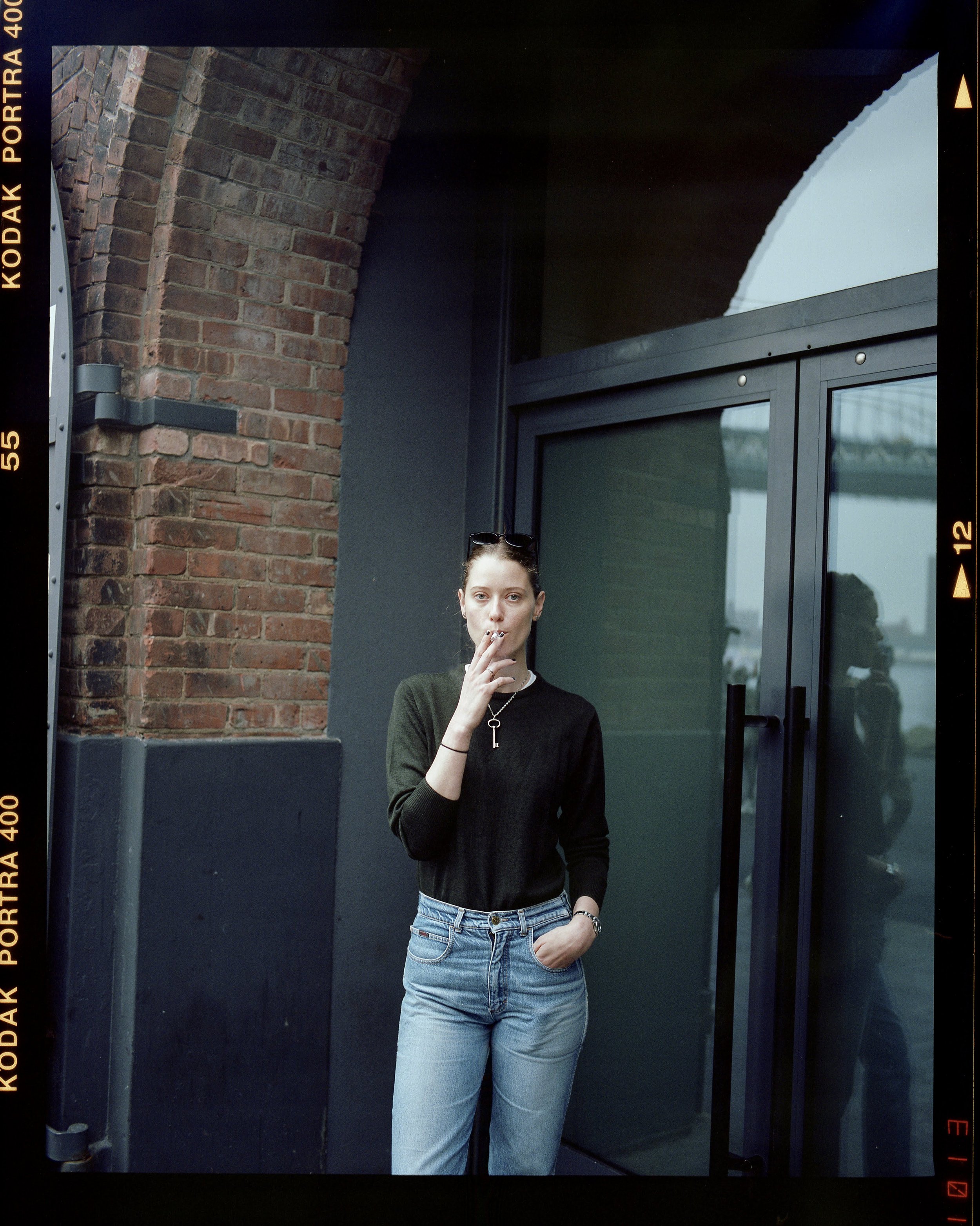 A young woman with sunglasses on her head, wearing a black sweater and light blue jeans, is standing outside near a brick wall and glass door. She is smoking a cigarette and looking at the camera.