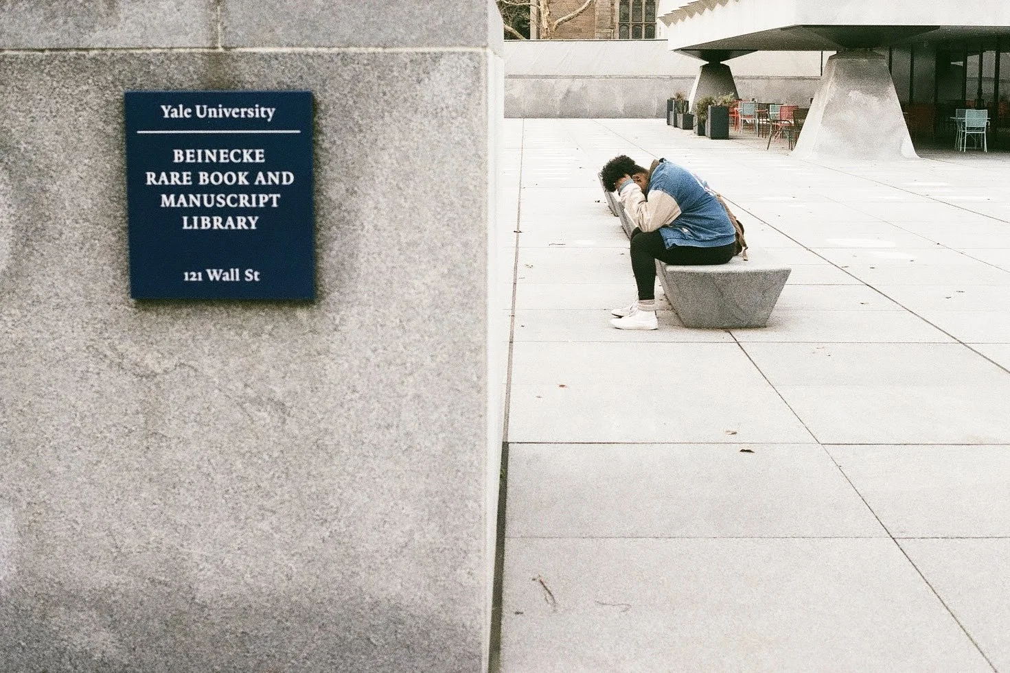 A person sitting on a concrete bench outside Yale University's Beinecke Rare Book and Manuscript Library, reading or studying with their head resting on their hand.