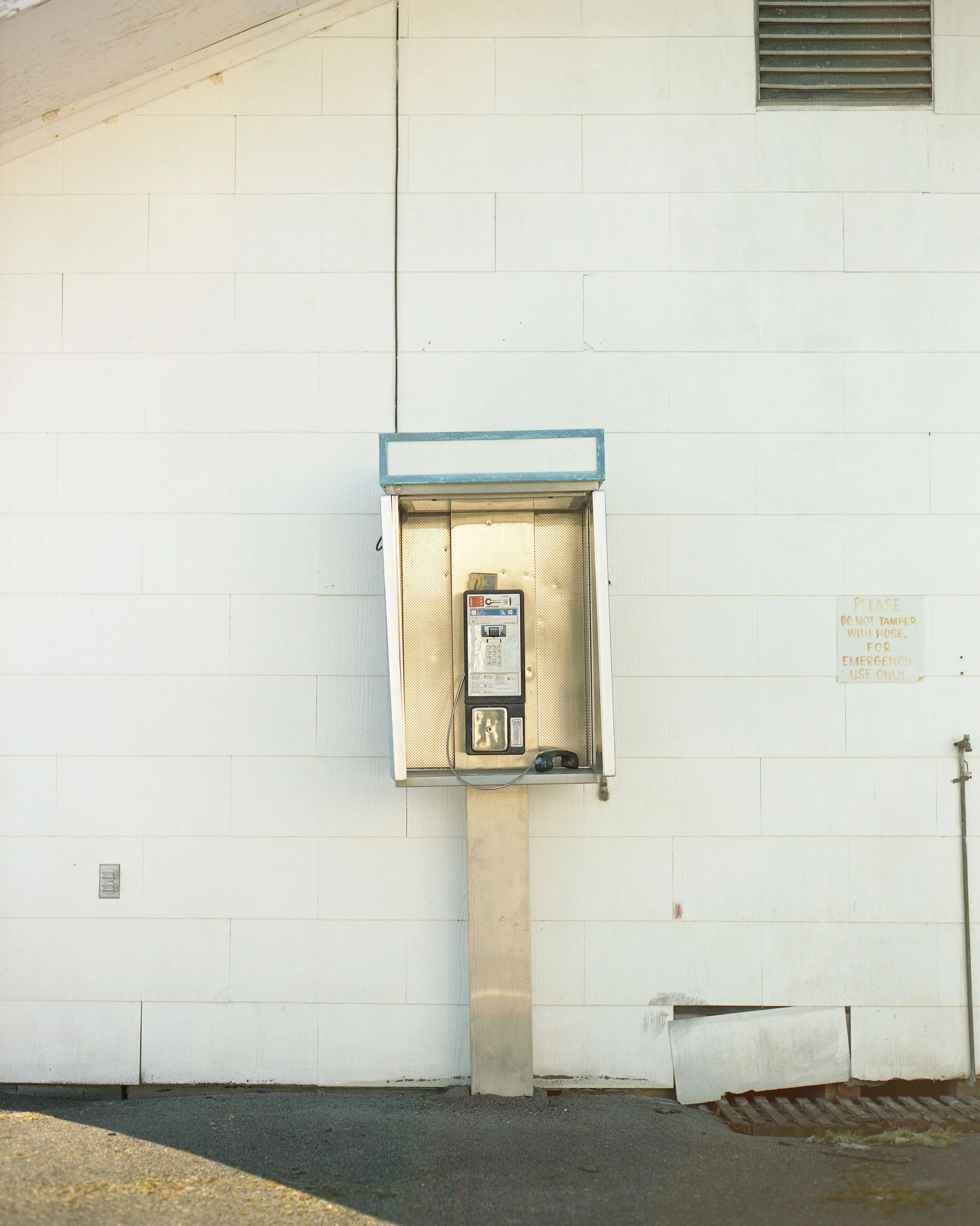 Public payphone mounted on a metal pole against a white wall. There is a small sign on the wall with instructions for emergency use.
