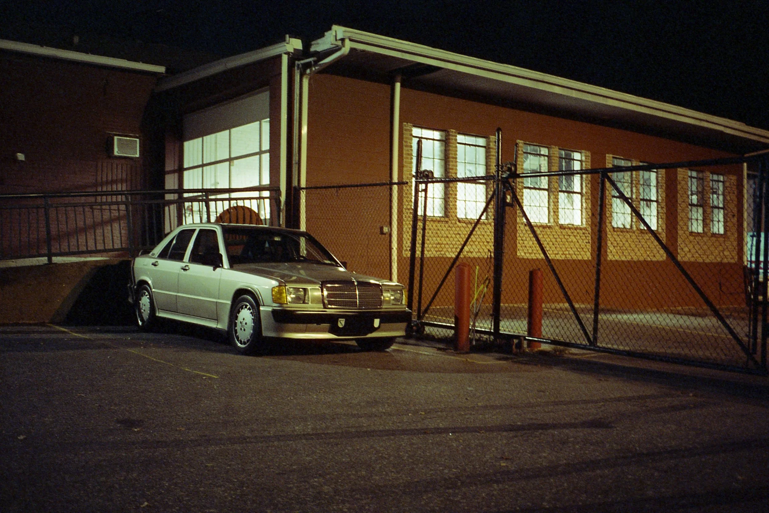 A silver vintage sedan parked on the side of a dark street at night, in front of a brick building with lit windows, surrounded by a chain-link fence and a parking lot.