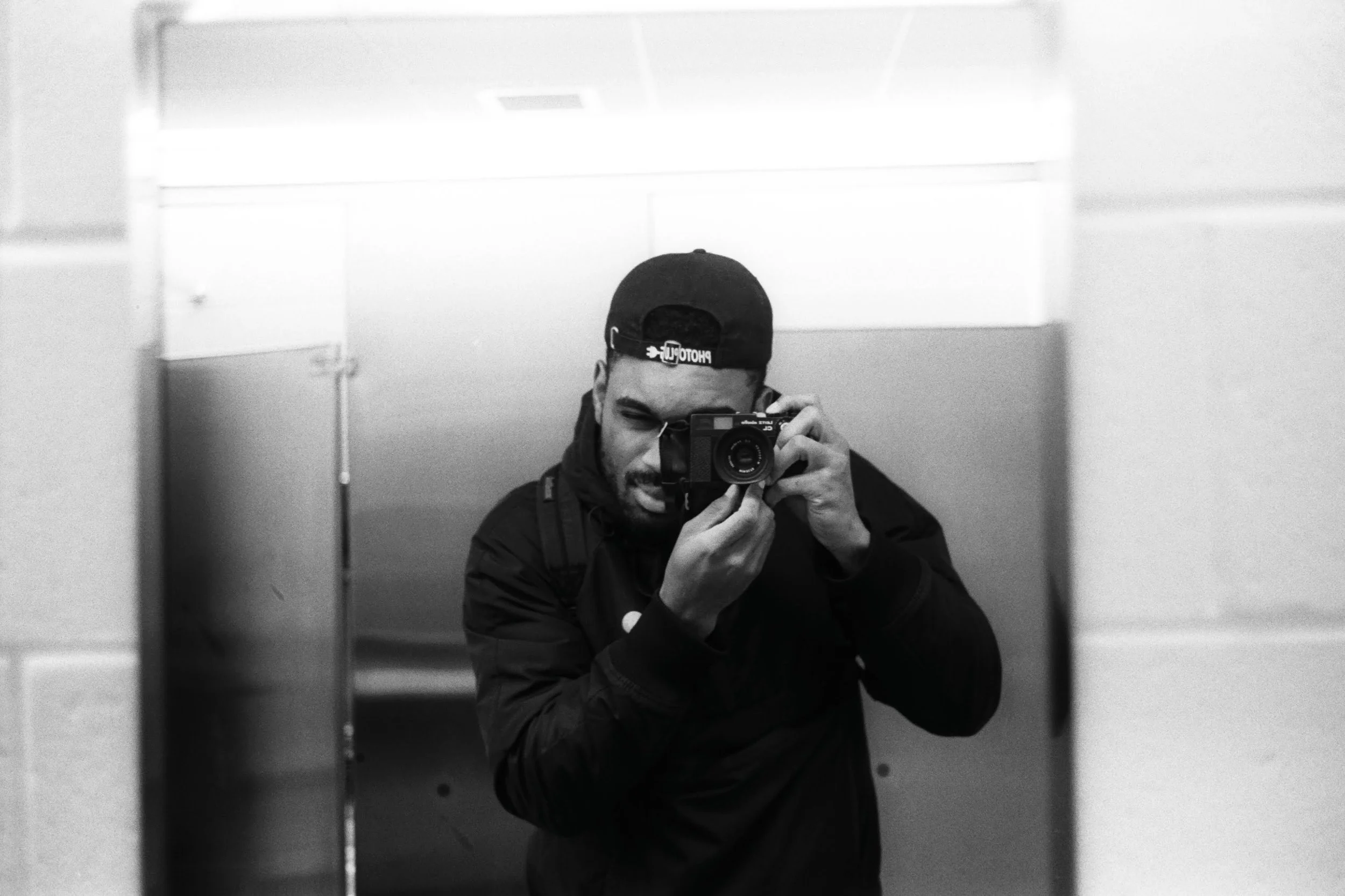 A person taking a selfie in a mirror with a camera, wearing a black jacket and baseball cap, in a restroom with tiled walls.