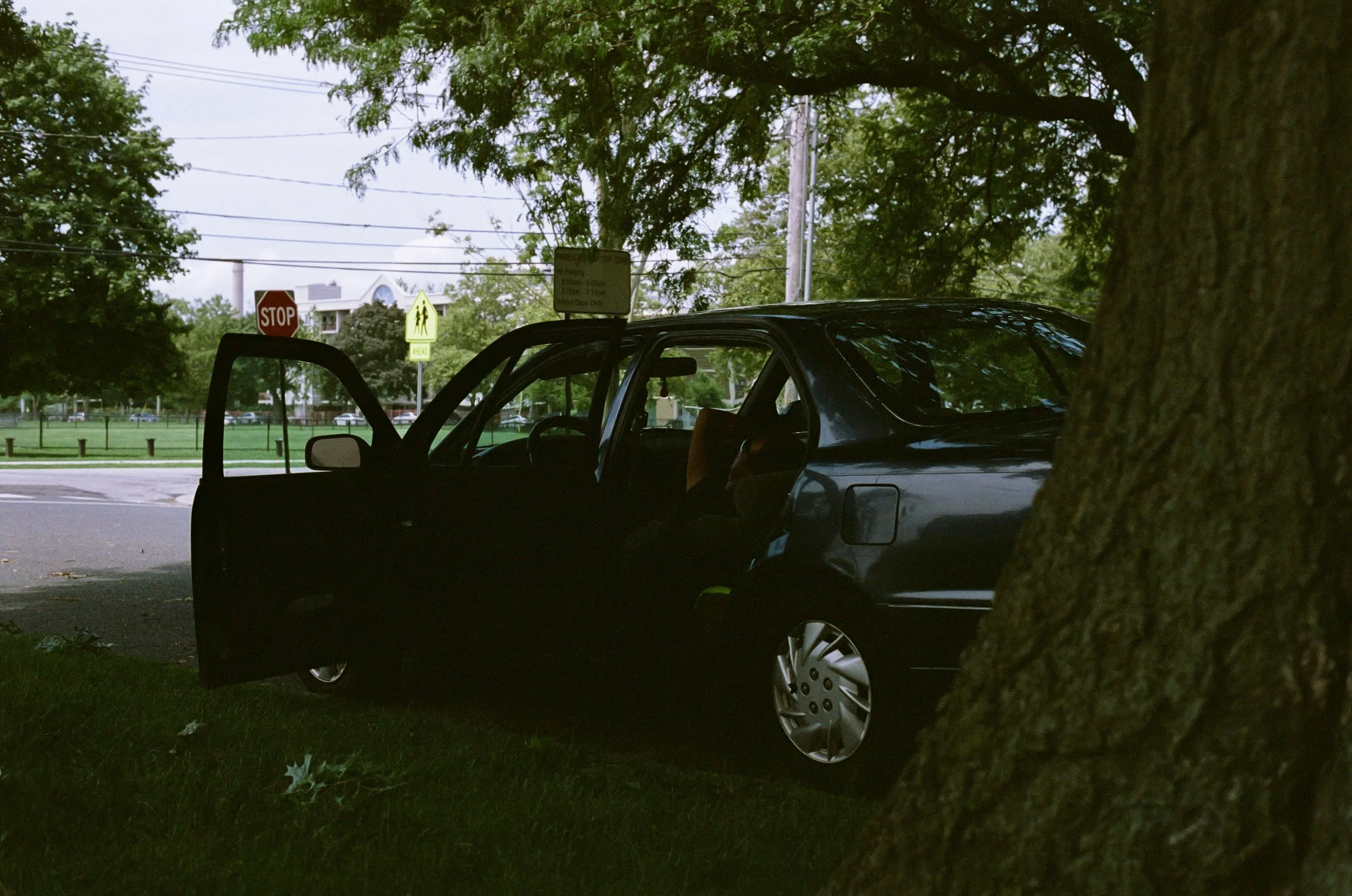 A dark-colored car parked on a grassy area under a tree, with its driver's side door open and a child sitting in the back seat.