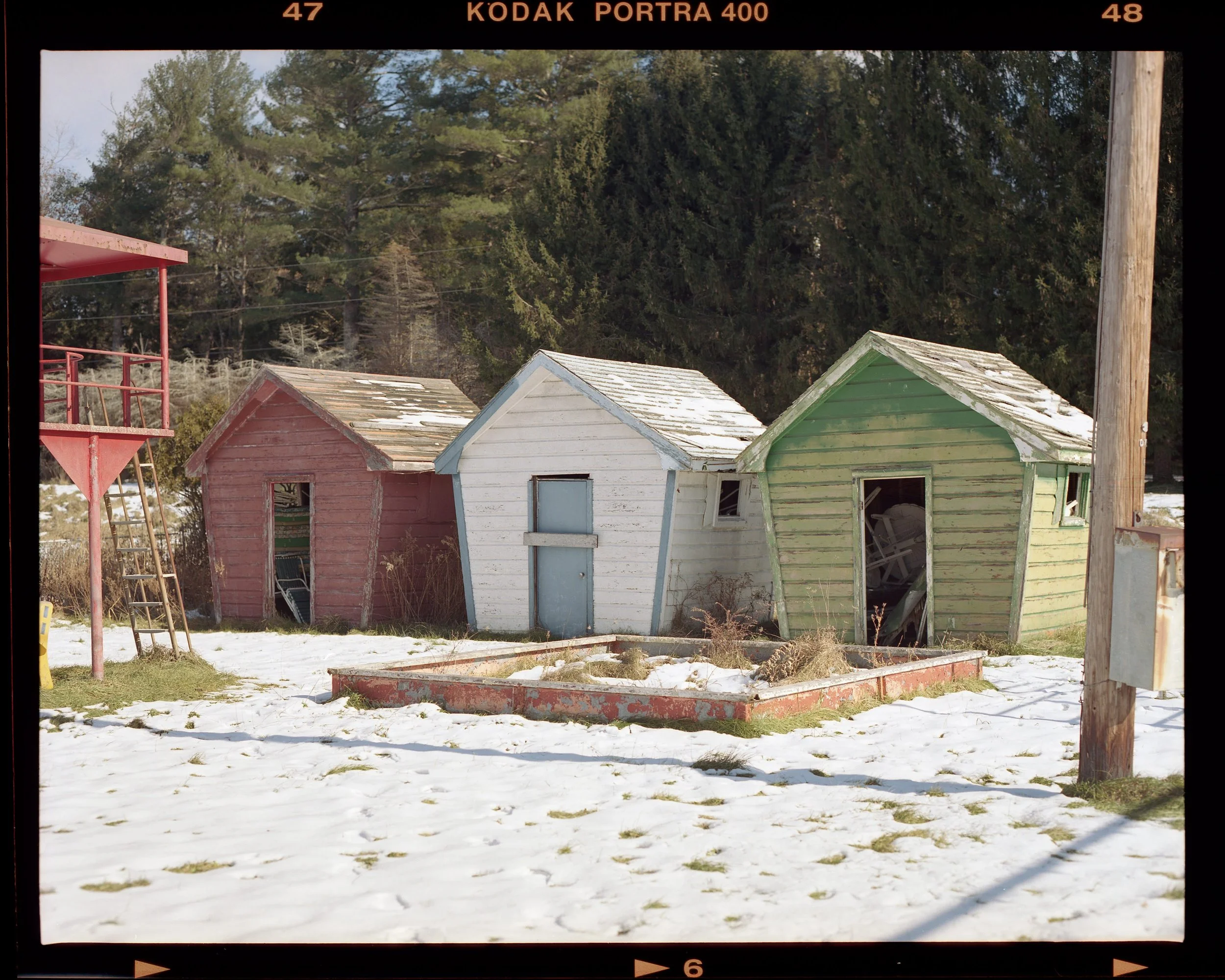 Three small, colorful, weathered wooden sheds in a snowy outdoor setting with trees in the background.
