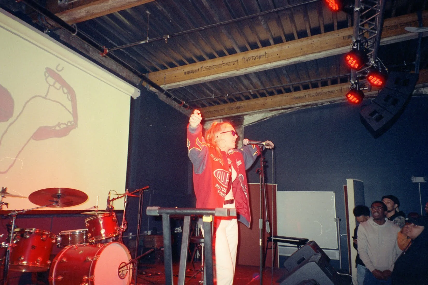 Performer with red hair and sunglasses speaking or singing into a microphone on a stage, with a band setup including a red drum set, and an audience watching in a dimly-lit venue.