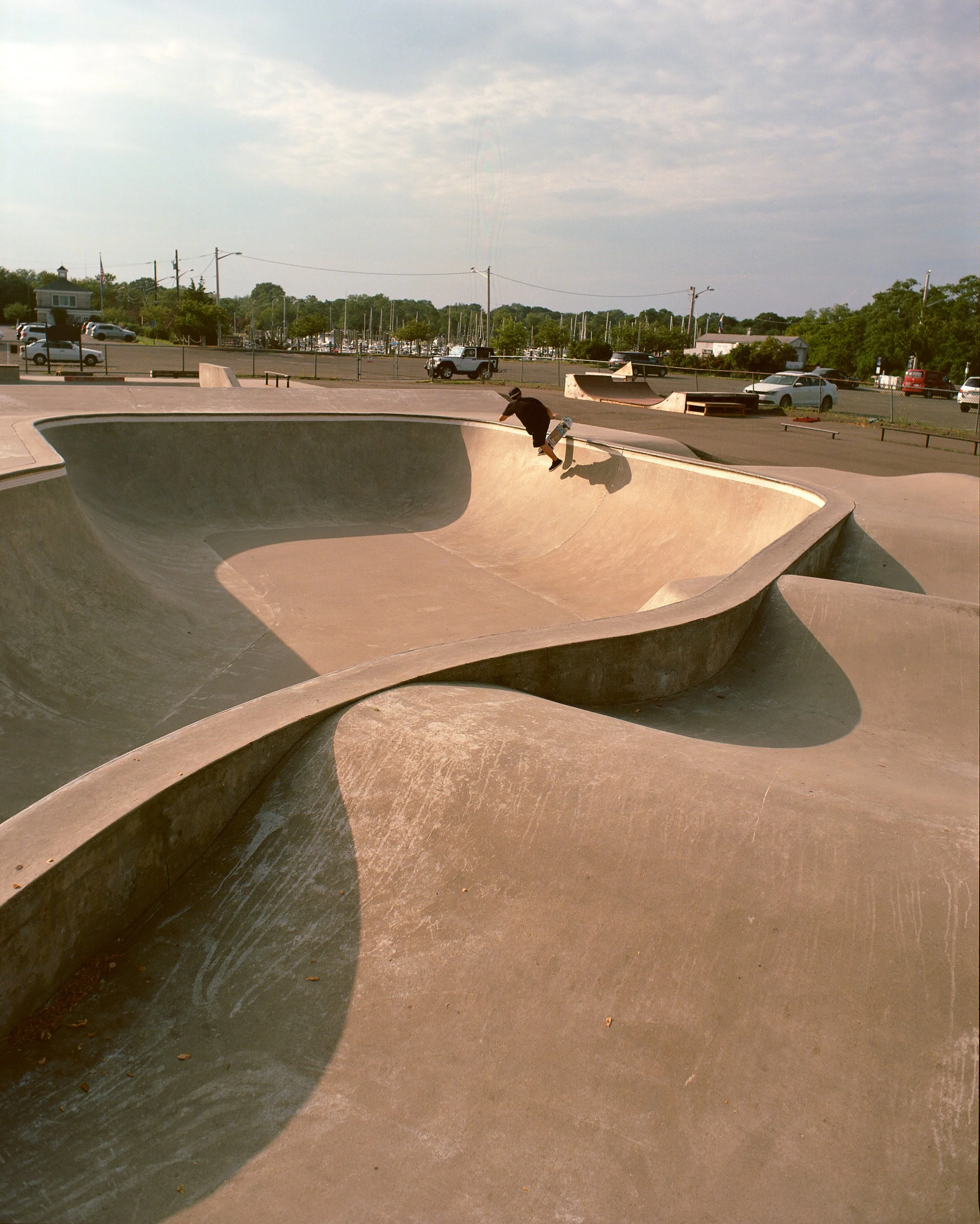 A skateboarder performing a trick in a concrete skate park with bowls and ramps, under a partly cloudy sky.