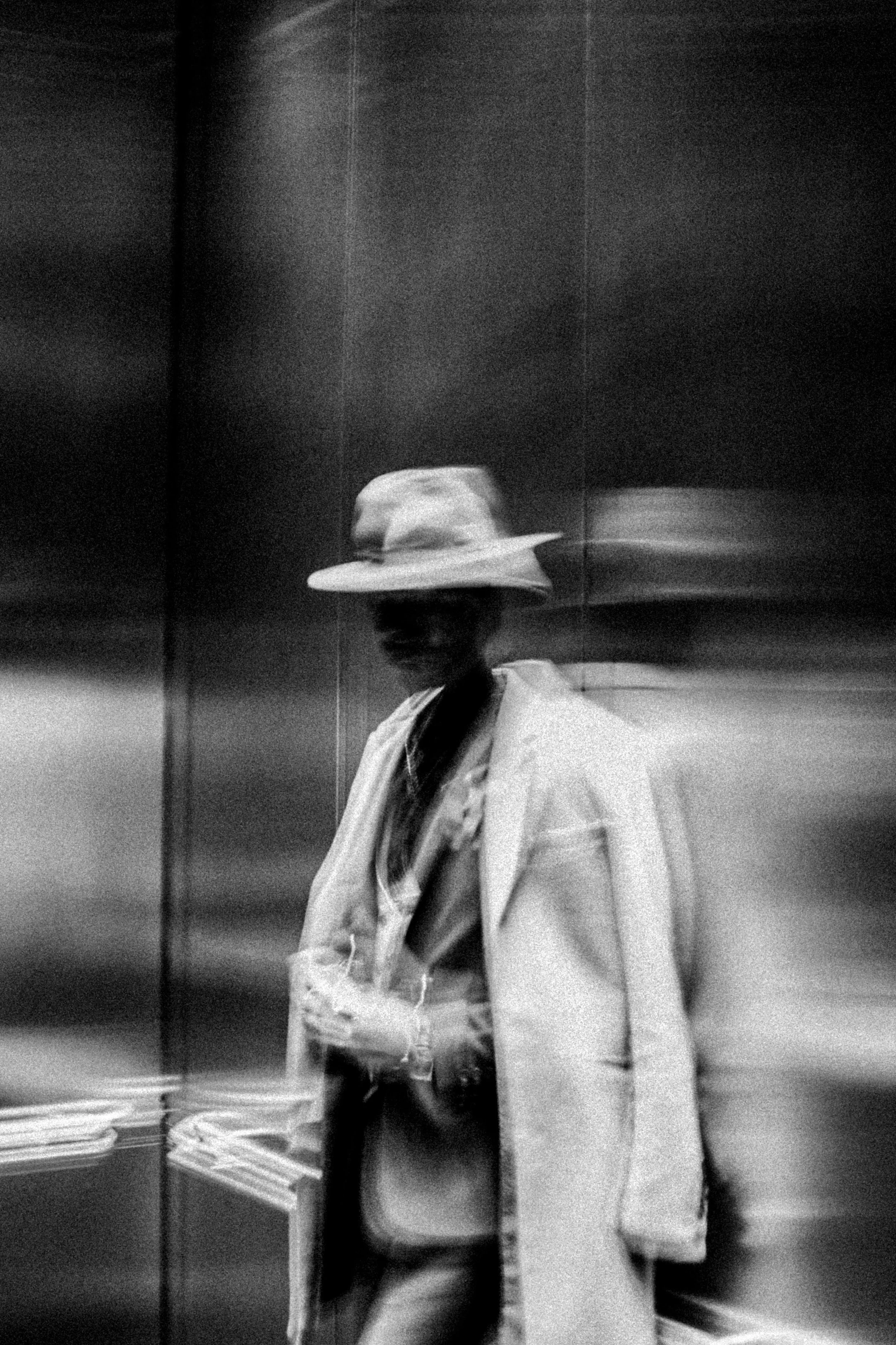 Black and white photo of a person wearing a wide-brimmed hat, coat, and scarf, standing in an elevator with motion blur.