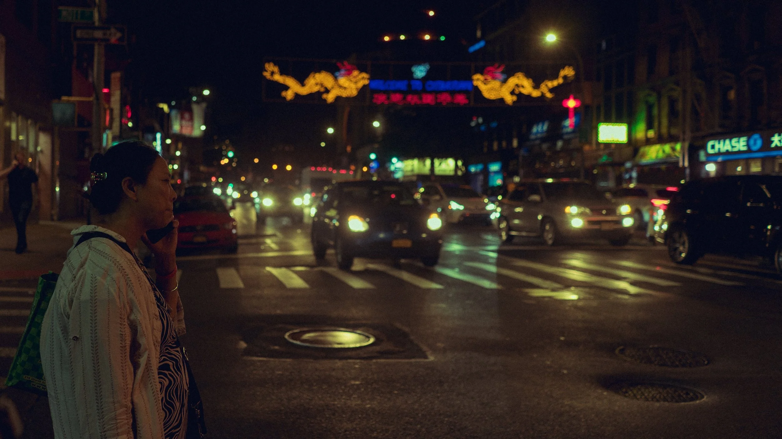A woman standing on the sidewalk talking on her cellphone at night, with a busy street and illuminated signs in the background.