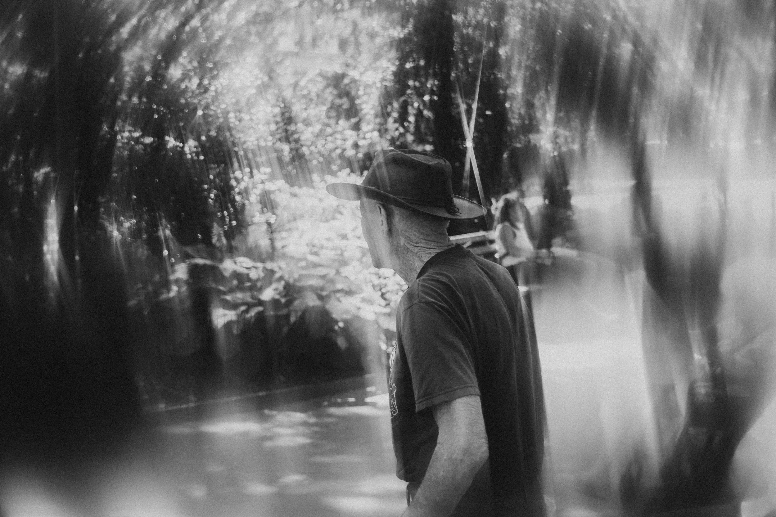 A black and white photo of a man wearing a hat, standing outdoors near a body of water with trees and other people in the background.