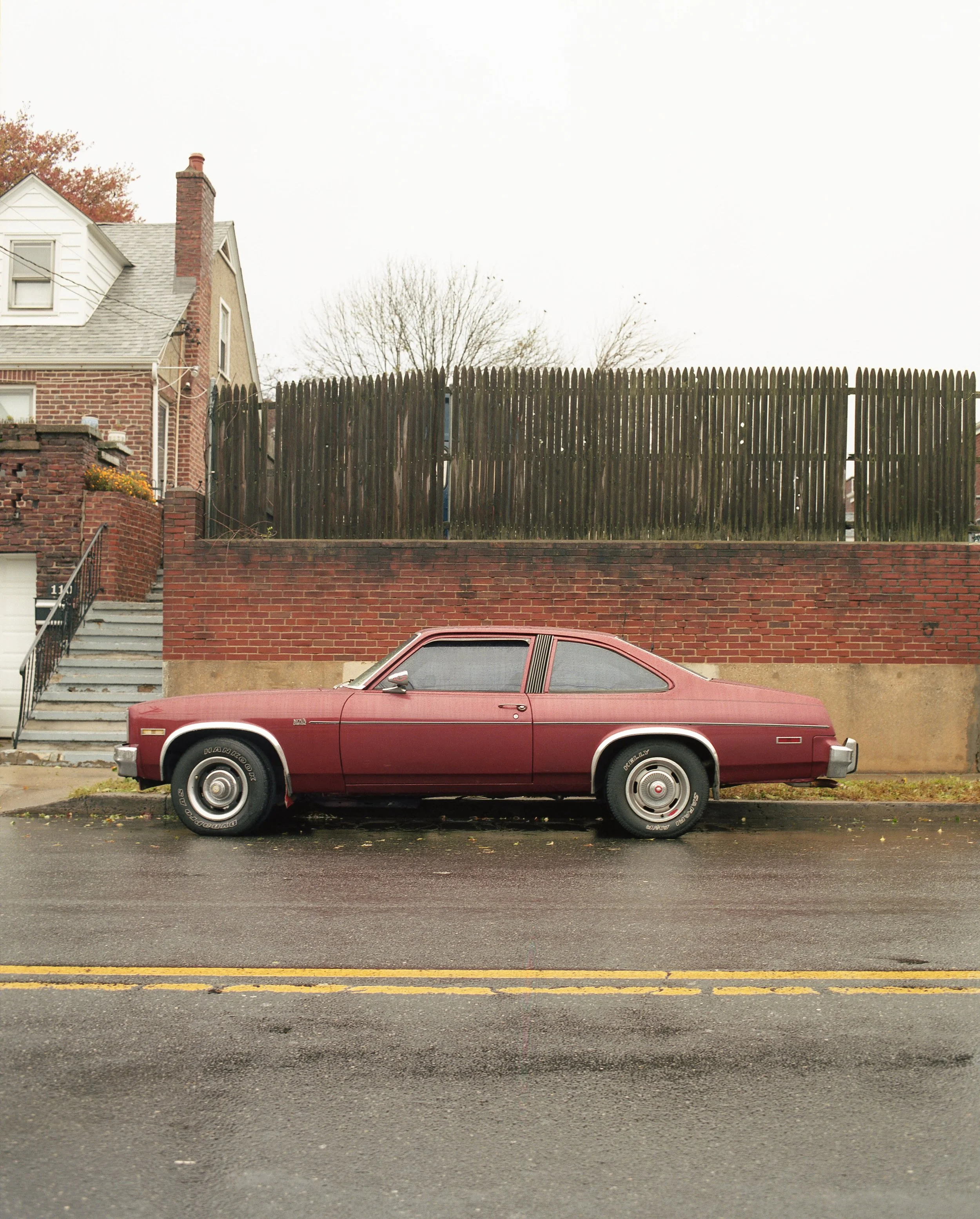 A vintage red two-door coupe car parked on the street in front of a brick wall and a wooden fence with a house and trees in the background.