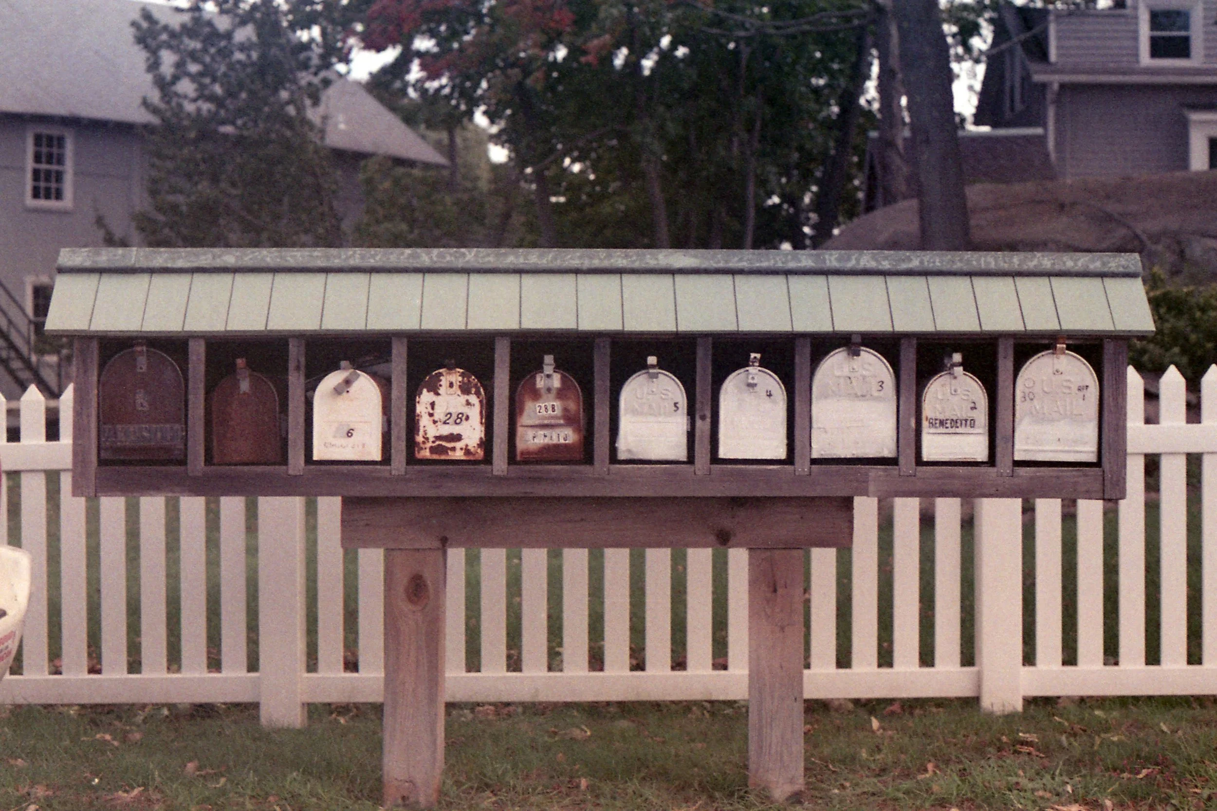 A row of old mailboxes mounted under a small roof on a wooden stand, with a white picket fence and houses in the background.