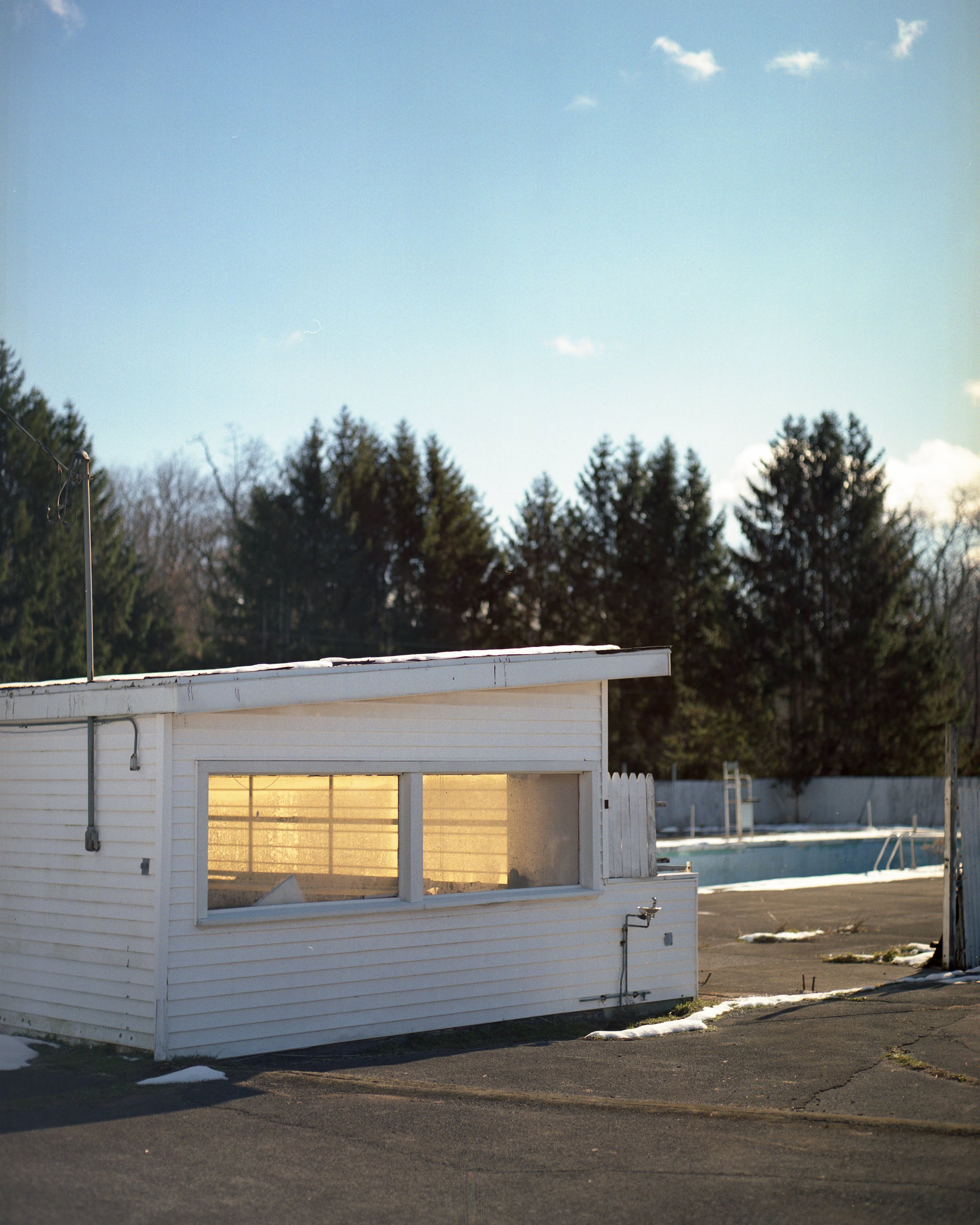 An empty outdoor swimming pool with a small white building nearby, surrounded by a concrete deck with some patches of snow, trees in the background, and a clear blue sky.