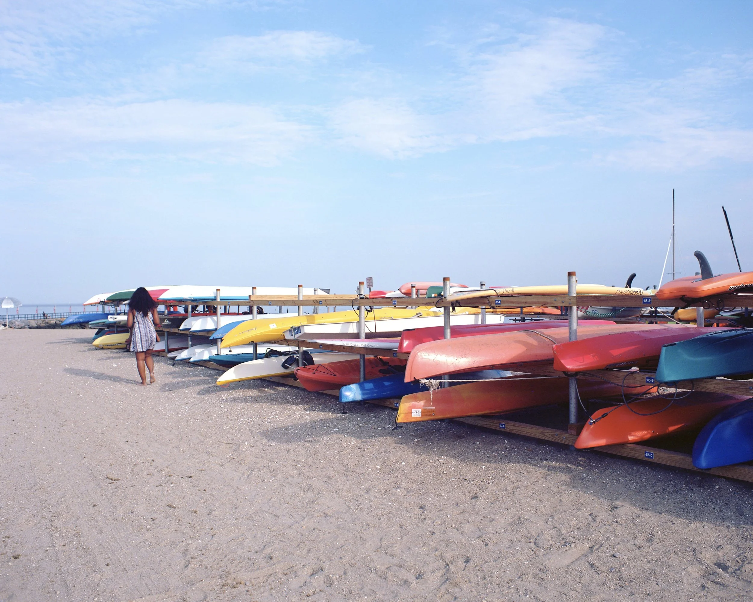Colorful kayaks and canoes stored on racks on a sandy beach, with a woman walking nearby and a blue sky overhead.