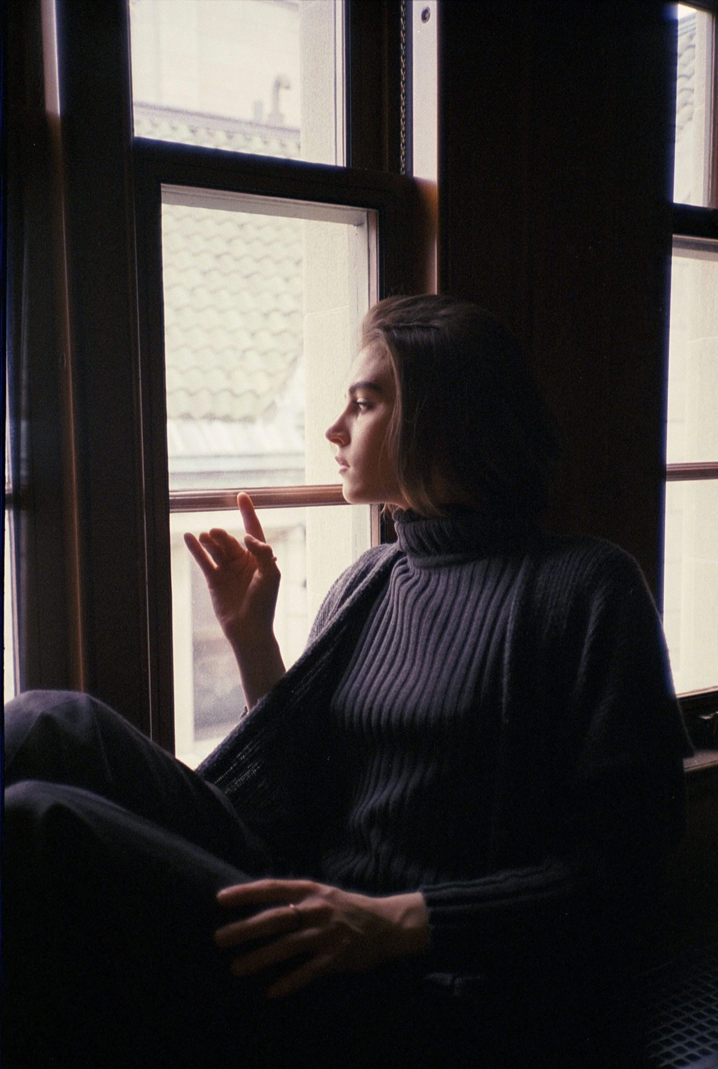 Young woman sitting by a window with daylight, wearing a dark ribbed turtleneck, looking outside with a thoughtful expression.
