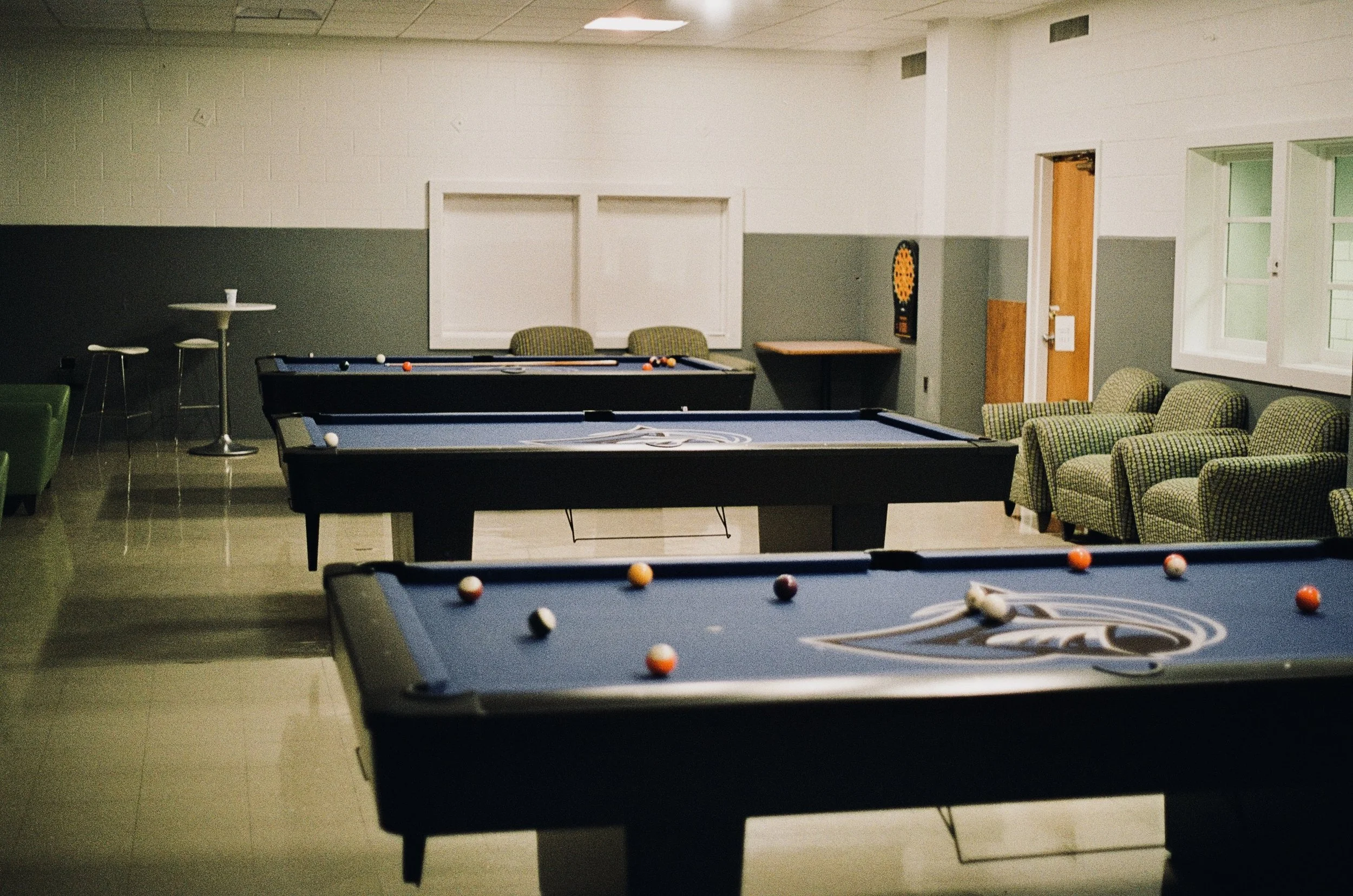 A recreational room with three pool tables, a dartboard, a small table with chairs, and green patterned armchairs near windows.