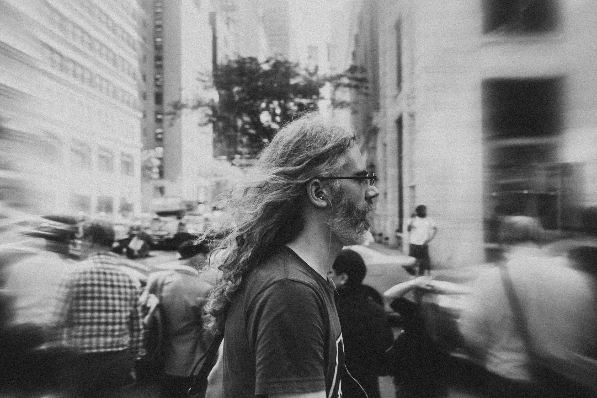 A black and white photo of a man with long, wavy hair and a beard, wearing glasses and earphones, walking on a busy city street with tall buildings and blurred pedestrians and vehicles in the background.